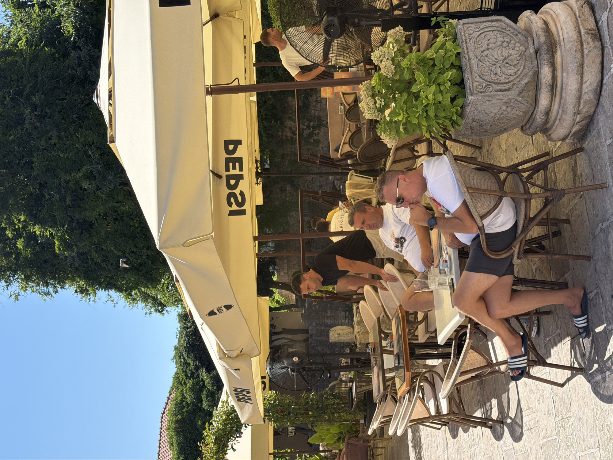 The image depicts an outdoor seating area of a cafe, likely in Budva, Montenegro, on a sunny day. The cafe is shaded by large, light-colored umbrellas with the "Pepsi" logo prominently displayed. Several people are seated at tables. In the foreground, a man wearing sunglasses and a white t-shirt is seated in a wicker chair, looking towards the right. Another man in a white t-shirt and dark shorts is sitting with his legs crossed, wearing dark flip-flops with white stripes. Behind them, a man in a black t-shirt stands near a table, looking to his left. Further back, another man in a white t-shirt is visible. The environment includes stone paving on the ground, green trees and foliage in the background, and large industrial fans for cooling. A decorative stone planter with a green plant is also present in the foreground. The overall atmosphere suggests a relaxed outdoor dining or lounging experience.