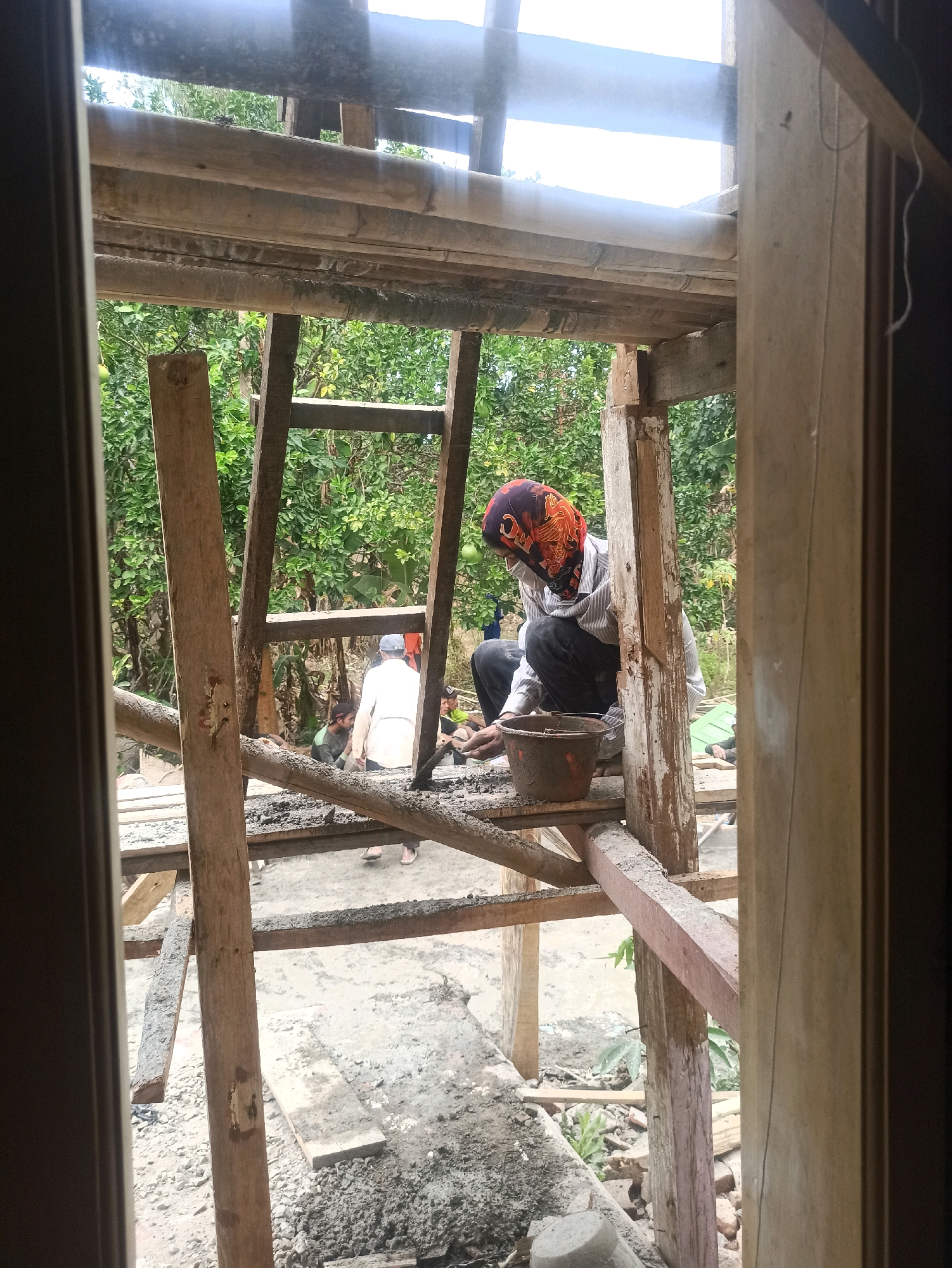 An exterior shot captures a construction site where several individuals are working on what appears to be a wooden structure. In the foreground, a person wearing a patterned headscarf, a light-colored striped shirt, and dark pants is crouched on a wooden beam, attending to a bucket filled with a grayish substance, possibly cement. Behind this individual, another person in a white garment and a blue cap is visible, along with other figures in the background amidst lush green foliage. The scene is characterized by exposed wooden beams, scaffolding, and debris scattered on the ground, suggesting ongoing construction. The lighting indicates it is daytime, with natural light illuminating the site.