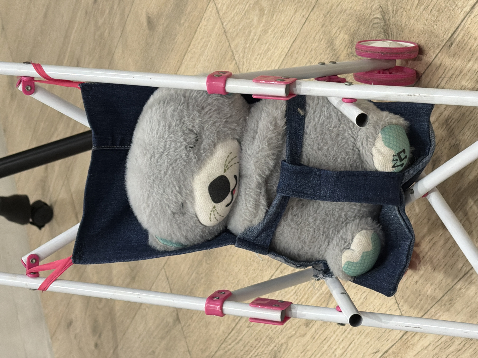 A close-up, high-angle shot shows a gray teddy bear with its eyes closed, sleeping in a miniature blue denim stroller. The stroller has a white metal frame and pink accents on its wheels and attachment points. The teddy bear is nestled in the denim seat, with its arms crossed. The background is a light-colored wooden floor. The image is well-lit and in focus. There is no information about the location of Krasnyi Krym, Russia in the image.