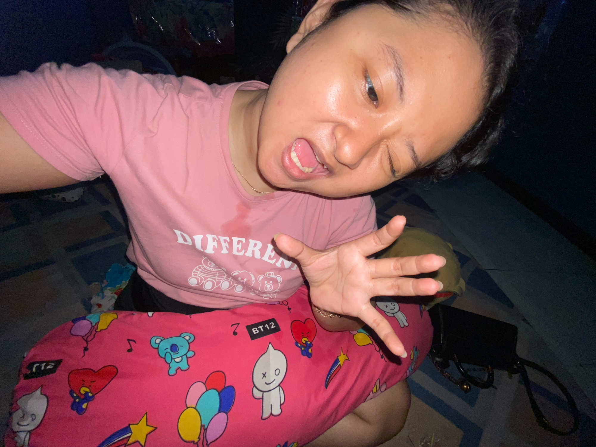 A woman is taking a selfie in what appears to be a residential setting. She is wearing a pink t-shirt with the word "DIFFERENT" printed in white, along with cartoon bear graphics. She is leaning back and has her mouth open, with her right eye partially closed. Her left hand is held up towards the camera, with fingers spread. She is resting on a pink pillow covered in cartoon characters and balloons. A black bag with a chain strap is visible in the background. The lighting suggests it is nighttime or indoors. There is no discernible information in the image that places the scene in Gelam, Indonesia.