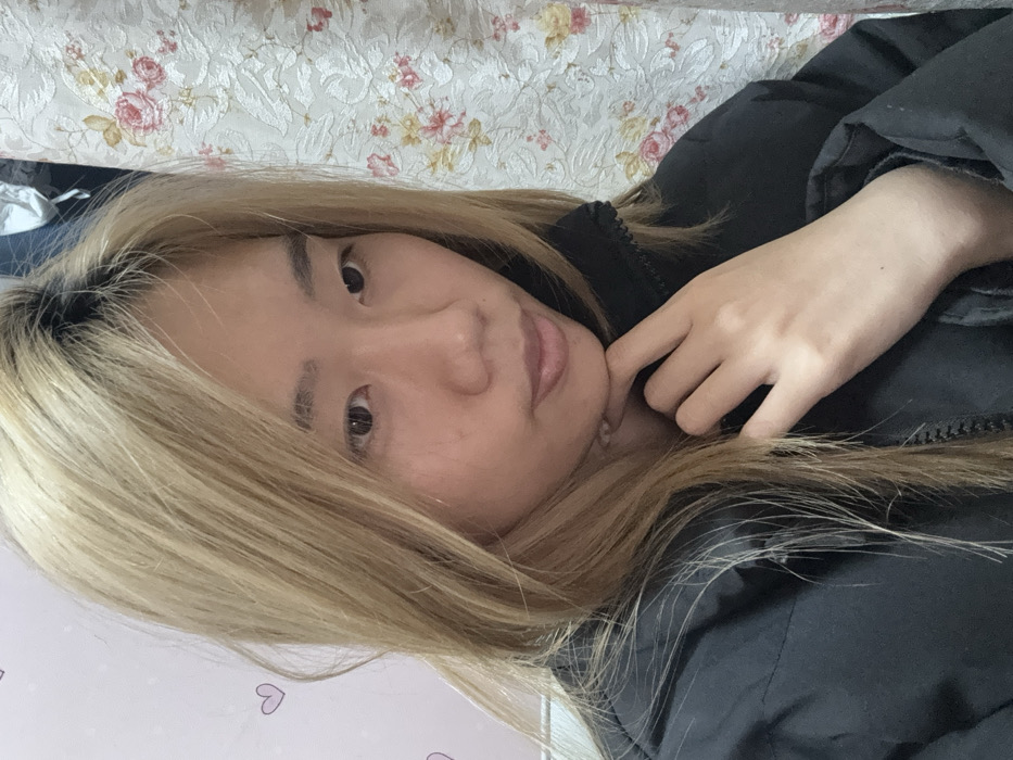 An East Asian female subject is depicted in a medium close-up, seated indoors within Shapingba District, China. The individual has long, straight, blonde hair with visible dark roots and is wearing a black puffer jacket with a zippered front and a ribbed collar. Her left hand is positioned with fingers slightly curled under her chin. She is looking directly towards the viewer with a neutral facial expression. The background on the left features a light-colored wall with small, pink heart outlines. On the right, a floral patterned curtain or wallpaper is visible, displaying large pink and red roses with green foliage on a light-colored, textured surface. A white garment or object is partially visible hanging in the upper right corner.
