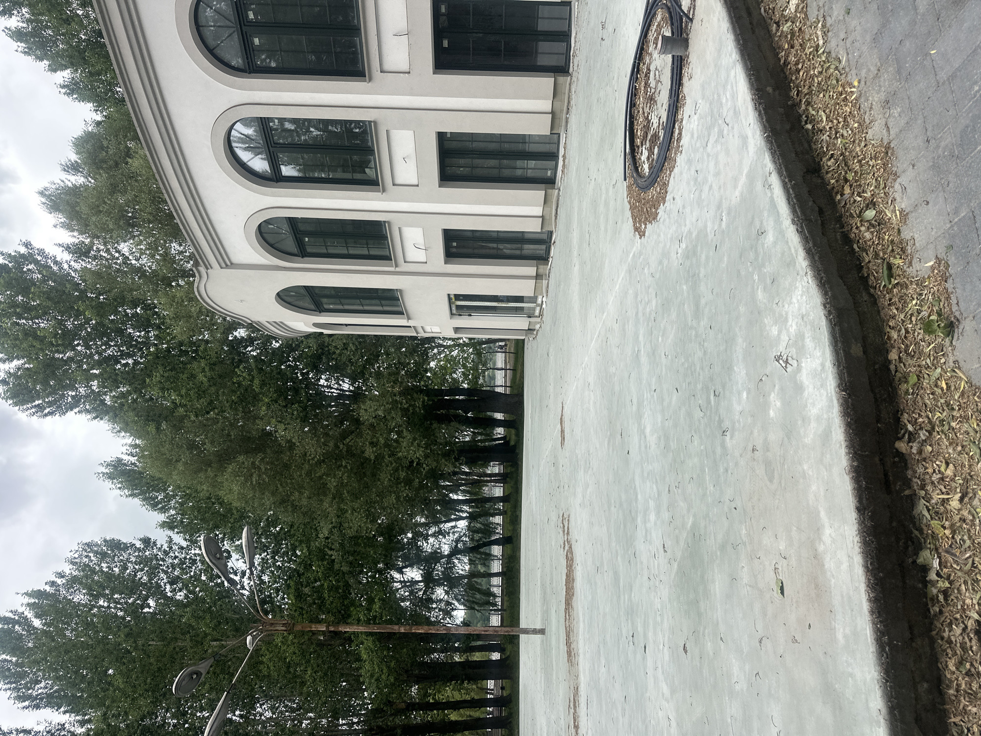 The image captures an exterior scene featuring a large, light-colored, modern building with arched windows and doors, situated on the right side. In front of the building is a wide, pale green paved area that appears to be a public space or plaza. The area is sparsely littered with dry leaves and small debris. To the left, a tall, weathered lamp post with multiple lights stands prominently. Behind the paved area and the building, a dense line of tall, green trees forms a backdrop. In the distance, through a break in the trees, a body of water and a decorative fence are visible. The sky is overcast and gray. A coiled black hose and a pipe are present on the paved surface to the right of the image. The scene suggests a park-like setting, possibly near a waterfront, within the city of Ternopil, Ukraine. No people are visible in the image.