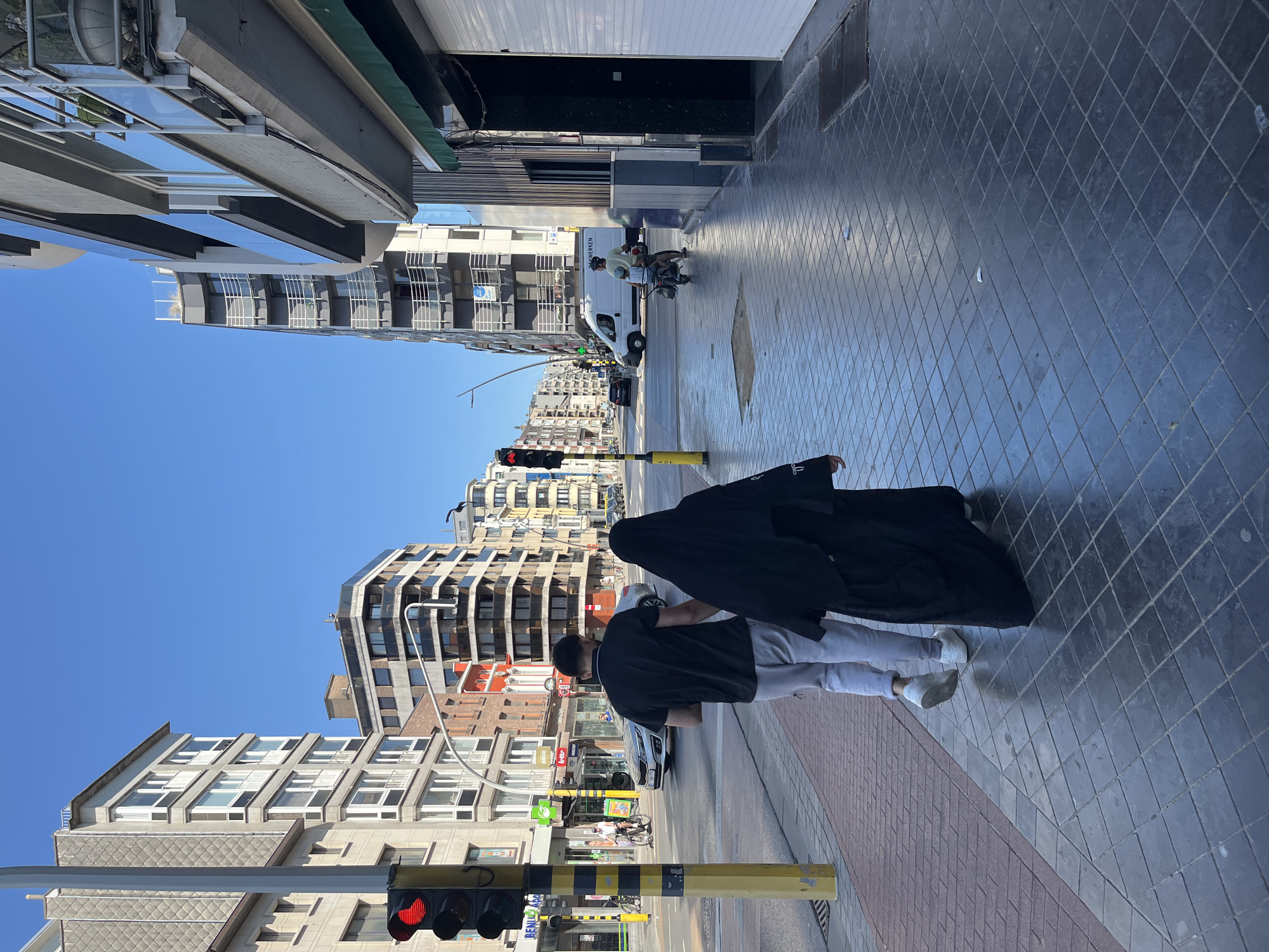 In Ostend, Belgium, on a sunny day, two individuals walk side-by-side along a tiled sidewalk. The person on the left, a male, wears a black t-shirt and light gray sweatpants, with white sneakers. The person on the right, a female, is dressed in a black abaya and hijab. They are walking away from the camera, on a sidewalk bordering a street with vehicles and buildings. A traffic light, showing red, is visible to the left of the frame. In the background, several multi-story buildings line the street, with a white van and a person on a moped also present in the scene. The ground is a mix of brick and dark square tiles.