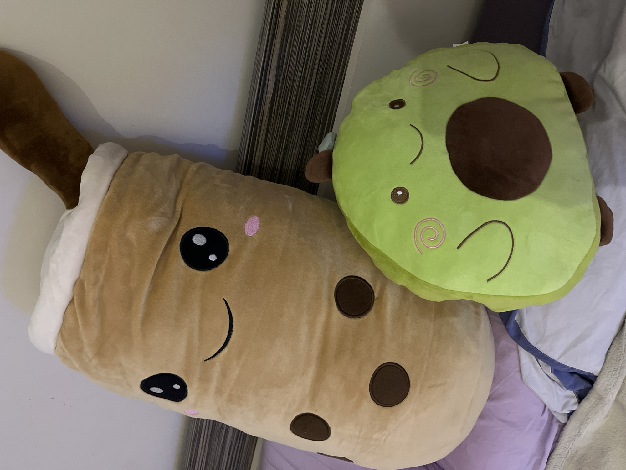 The image shows two plush pillows, one shaped like a bubble tea drink and the other like an avocado. The bubble tea pillow is brown with black eyes and a smile, and has brown circles representing the tapioca balls. The avocado pillow is green with a brown pit and smiling face. The pillows appear to be resting on a bed with purple and white sheets. Part of a headboard is visible in the background. The specific location in Veliko Tarnovo, Bulgaria, cannot be determined from the image.