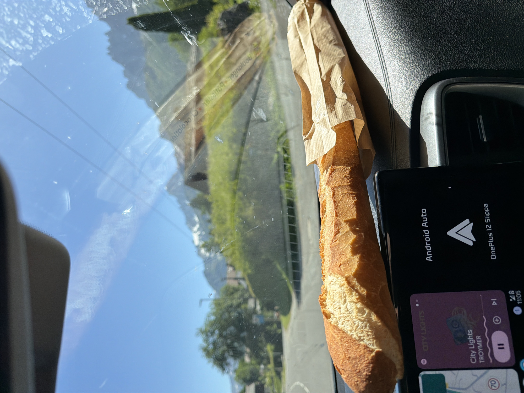 A close-up, forward-facing, eye-level shot captures a baguette resting on a car's dashboard. The bread, a light brown crust with a white interior, is partially wrapped in a brown paper bag and extends across the frame from left to right. Below the baguette, a car's infotainment screen displays "Android Auto" and music information for "City Lights" by Troymar. The background, seen through the windshield, reveals a bright blue sky with some clouds, lush green trees, houses, and snow-capped mountains, consistent with the location of Les Houches, France. The scene is illuminated by natural daylight.
