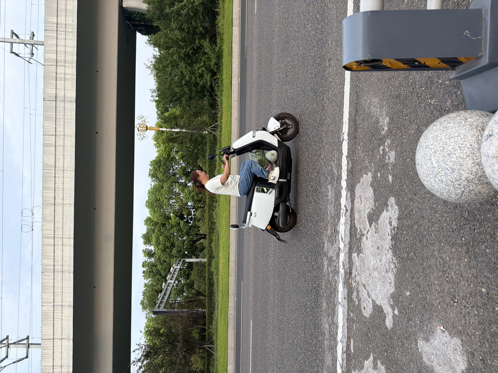 A woman is riding a white electric scooter down a paved road in Jiangjin, China. She is wearing a white short-sleeved shirt and blue jeans. The scooter has a license plate, some bags of produce, and a decorative sticker on the side. The road is lined with trees and a grassy median. An elevated concrete structure, likely a bridge or overpass, is visible in the background, with power lines running overhead. The sky is overcast. In the foreground, a guardrail with yellow and black stripes and decorative stone spheres are visible.