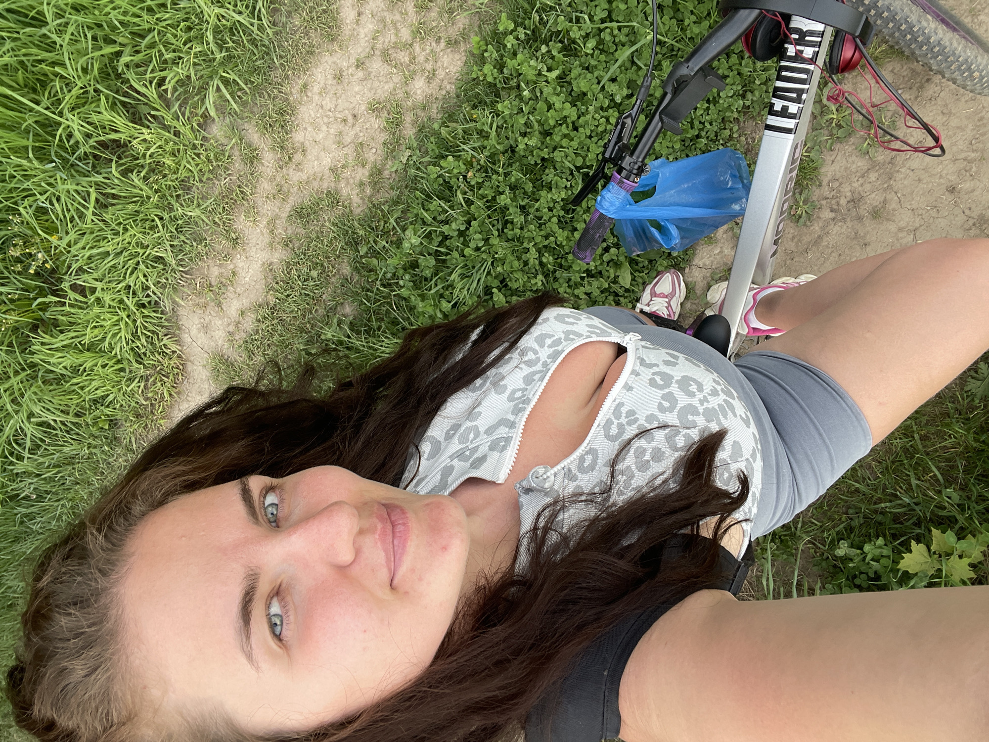 A woman is positioned at the bottom of the frame, facing the camera with her head tilted. Her expression is neutral, and she has long, dark hair. She is wearing a light-colored top with a white and grey print and a low zipper, grey shorts and dark top. Beside her is a bicycle; the bicycle frame is light-colored with the word "LEADER" visible. A pair of red headphones rests on the bike's handlebars, and a blue plastic bag is hanging from the bike. The background of the image includes a patch of grass and a dirt path. The location is outdoors and seems to be a grassy area near a dirt trail. This scene does not provide enough detail to identify this location as Ostrov, Russia.