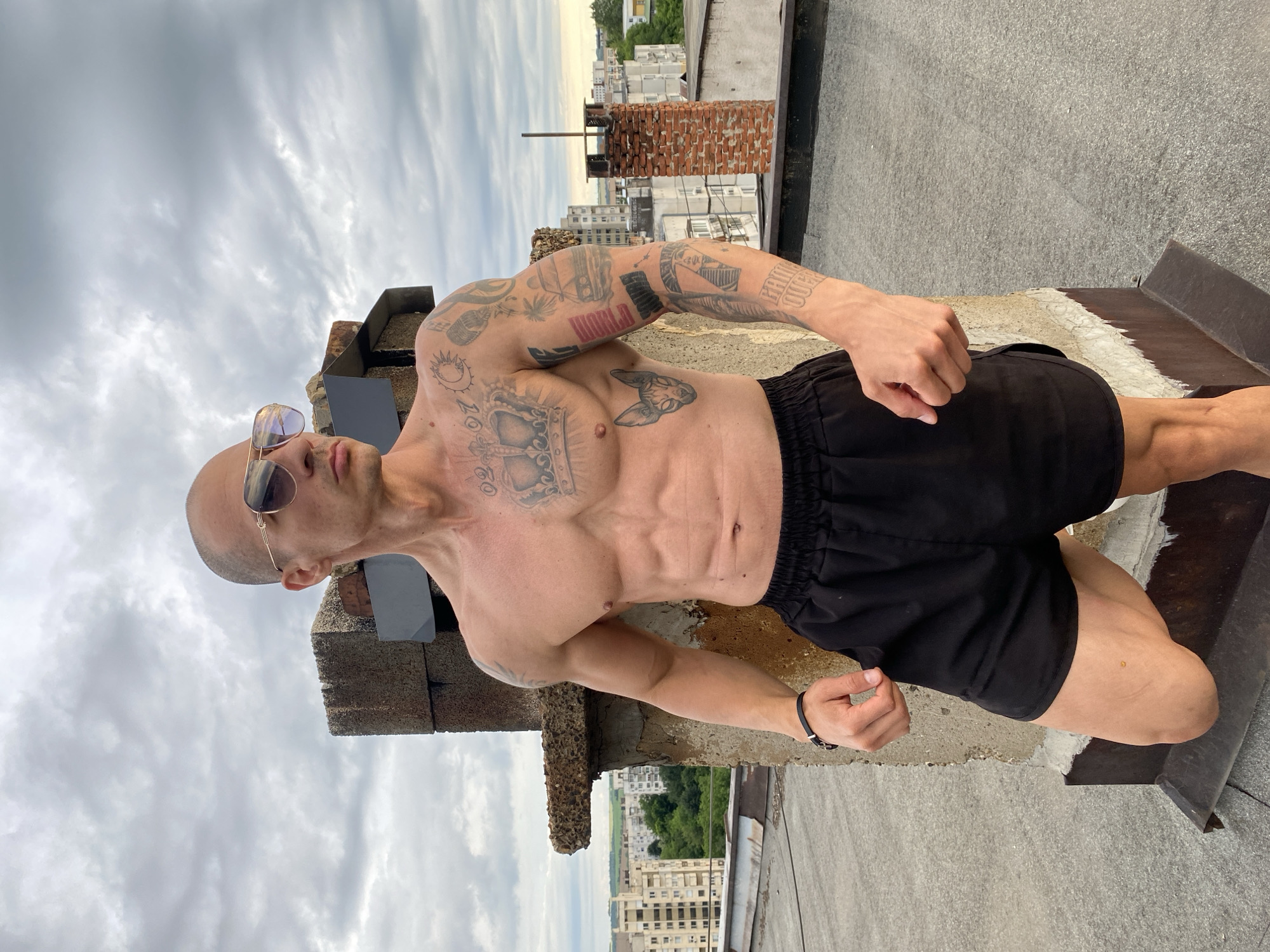 A bald, muscular man with tattoos on his chest and arms is standing on a rooftop. He is wearing aviator sunglasses and black shorts. He is looking to his right. In the background, there are buildings and a cloudy sky. The location is the city of Pleven, Bulgaria.