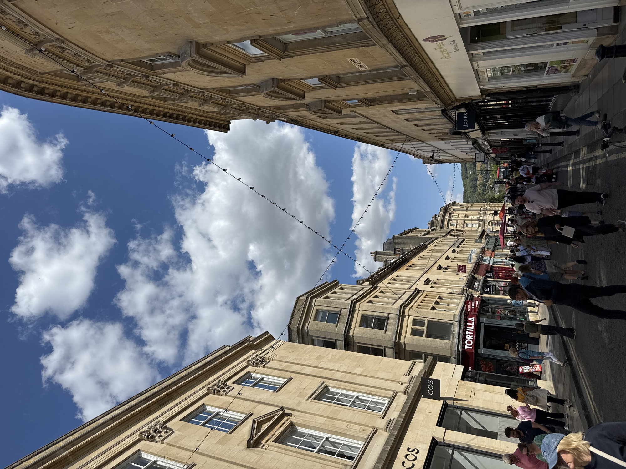 This is a ground-level, eye-level, medium shot photograph of a bustling street scene in Bath, United Kingdom, on a sunny day with a partly cloudy sky. The perspective is looking down a street lined with Georgian architecture, characterized by its honey-colored stone buildings.
On the left, the large building features several stories with uniformly spaced windows, some with decorative pediments. The ground floor has large commercial storefronts, including one with "COS" visible on the signage. On the right, the buildings also display Georgian architectural styles with arched windows and decorative cornices. A street sign reads "Cheap Street".
The street itself is paved with asphalt and marked with yellow lines. A crowd of people are walking in both directions, some appearing to be tourists. There are various shops and businesses lining the street, including "Tortilla," "Jones," and "Grape Tree" (a store selling "Eat Great Foods"). String lights are strung across the street between buildings. In the distance, rolling green hills are visible, suggesting the city's picturesque setting. A red double-decker bus is seen in the background. The overall impression is of a vibrant and historic urban environment.