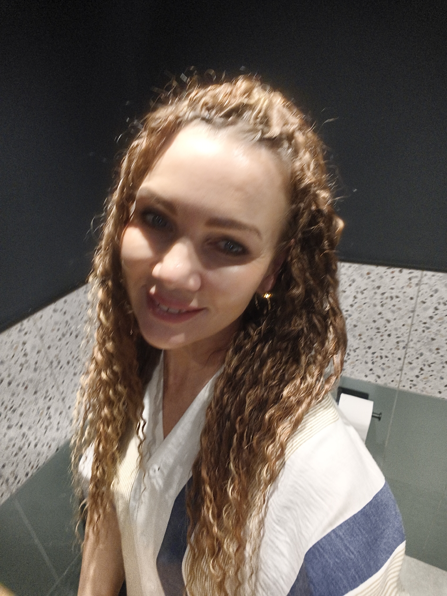 A woman with curly blonde hair and wearing a white and blue striped shirt is standing in a bathroom. The background consists of a speckled countertop and a dark wall. A roll of toilet paper is visible behind her shoulder. The location is Unknown, Unknown.