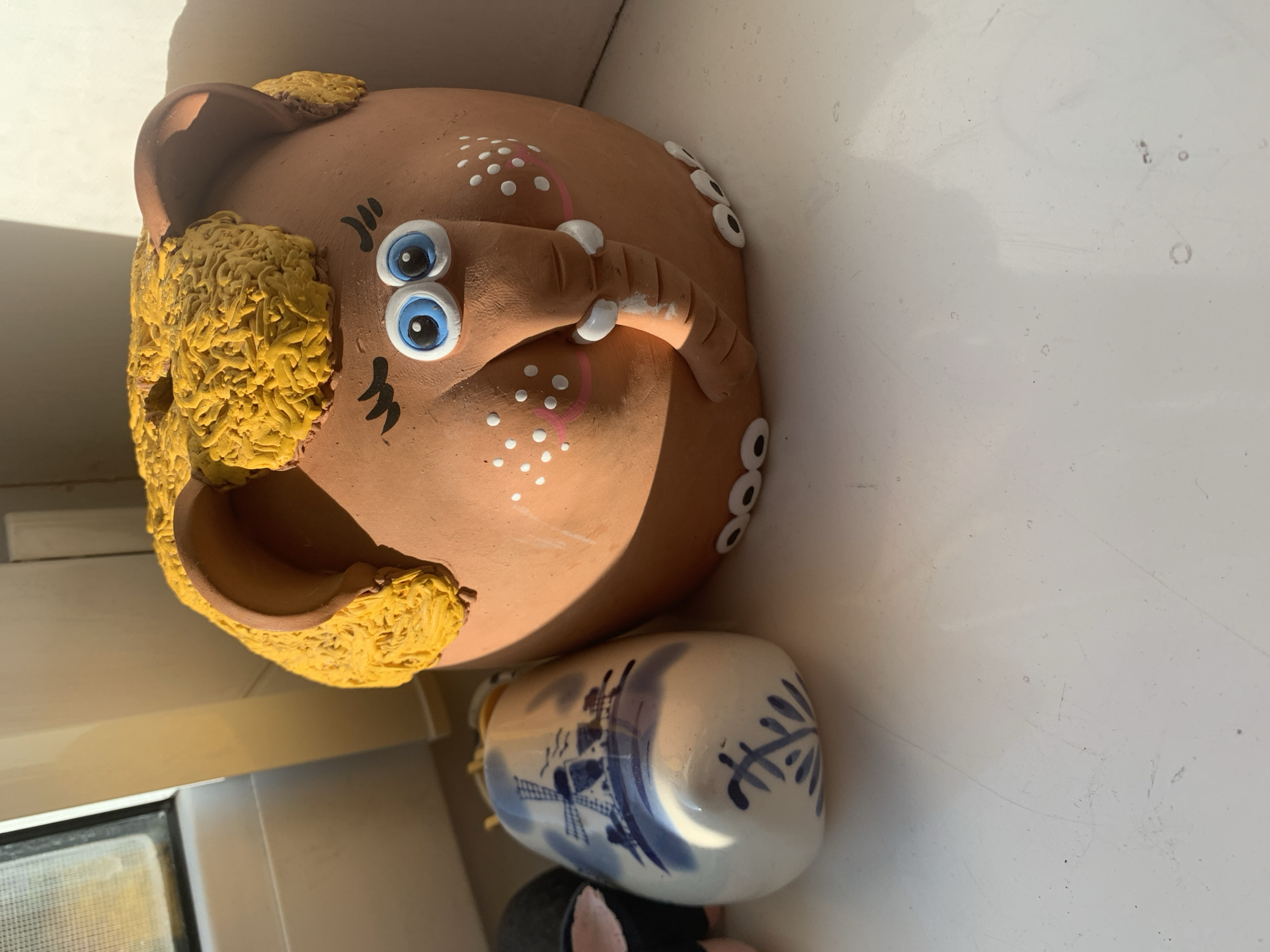 The image displays several decorative objects on a white surface, possibly a windowsill, given the presence of a window frame and natural light. The primary subject is a terracotta elephant figurine with large blue eyes, pink cheeks with white polka dots, and a bright yellow, textured "hair" or mane. Its trunk is curled downwards, with two white accents on it. To the left of the elephant is a white ceramic object decorated with blue Delft-style windmill and foliage patterns. Part of another dark object with pink accents is visible to the far left, possibly another figurine. The overall scene is brightly lit, with shadows casting from the objects and the window frame. There is no indication of the specific location within the city of Kapyĺ, Belarus.