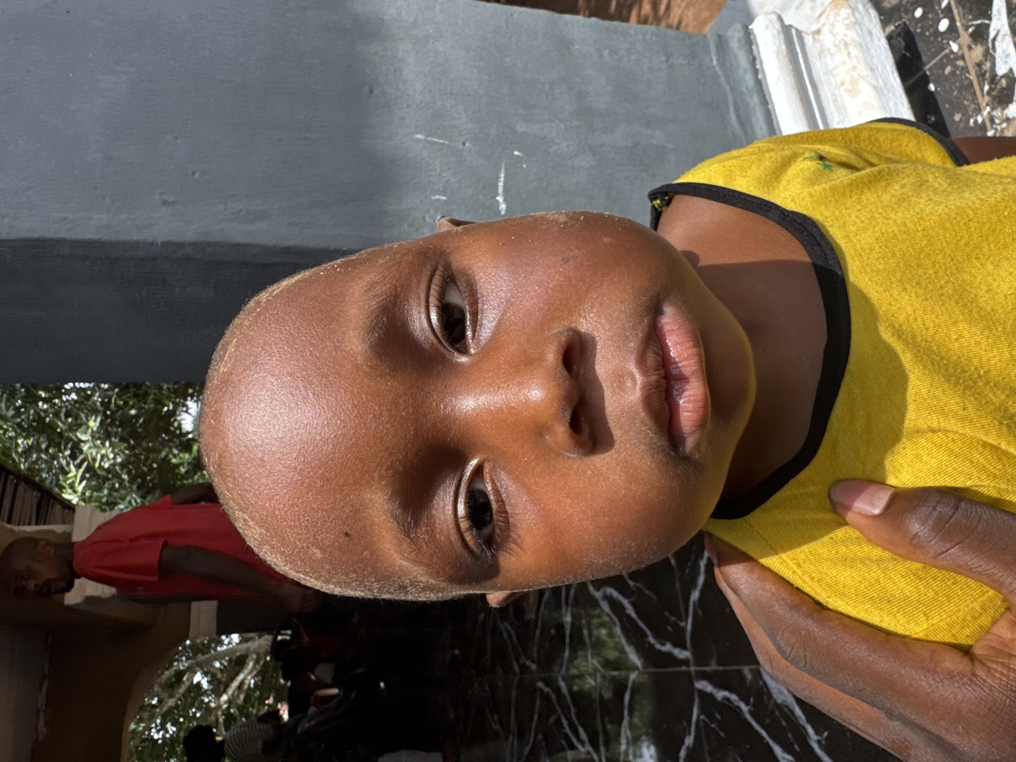 This close-up, portrait-style photograph, taken in Eziudo, Nigeria, features a young child with dark skin, facing directly towards the camera. The child's face, particularly the forehead and cheeks, is lightly covered in fine dust or sand. They are wearing a bright yellow t-shirt with black trim along the neckline and sleeves. An adult's hands, matching the child's skin tone, are visible in the lower right quadrant, appearing to gently hold or support the child.
The setting is outdoors during daylight, indicated by strong sunlight creating clear highlights and shadows on the subjects and environment. Behind the child's head is a grey, textured wall or structure. In the lower right, a dark, reflective surface, possibly tiled or marbled, shows bright reflections. In the background on the left, a person wearing a red shirt is partially visible, standing near green foliage, suggesting trees or bushes. The overall atmosphere is natural and informal. The child's expression is neutral and direct. There is no visible text in the image.
