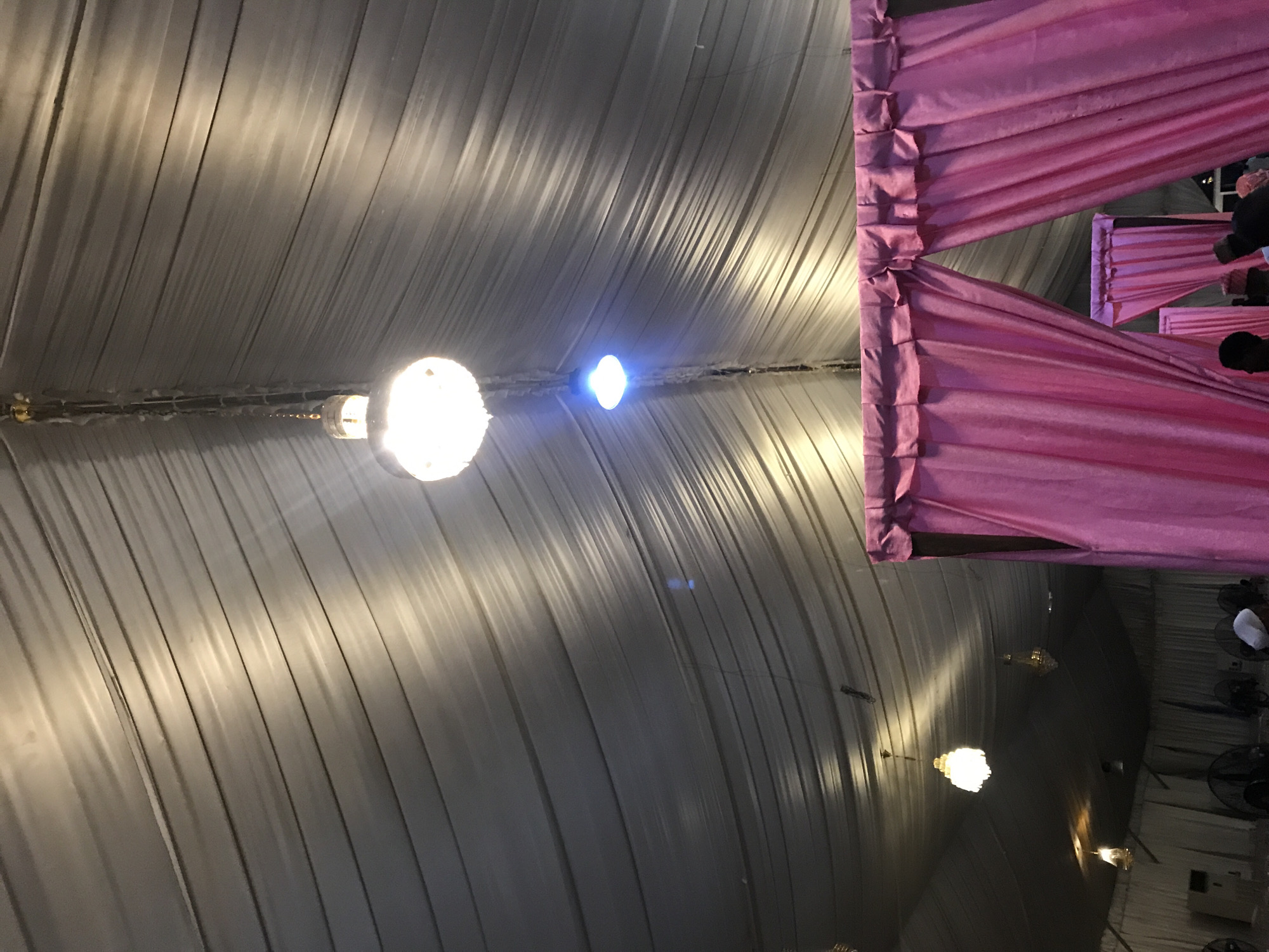 The image shows the interior of a large tent structure in Oko Erin, Nigeria. The tent has a gray, fabric ceiling with visible seams and folds. A string of lights hangs across the ceiling, with one bulb brightly illuminated in the center of the frame and another emitting a blue glow nearby. Pink fabric drapes decorate the sides of the tent. In the background, a dark space is visible, with hints of people and objects like fans and electrical equipment.