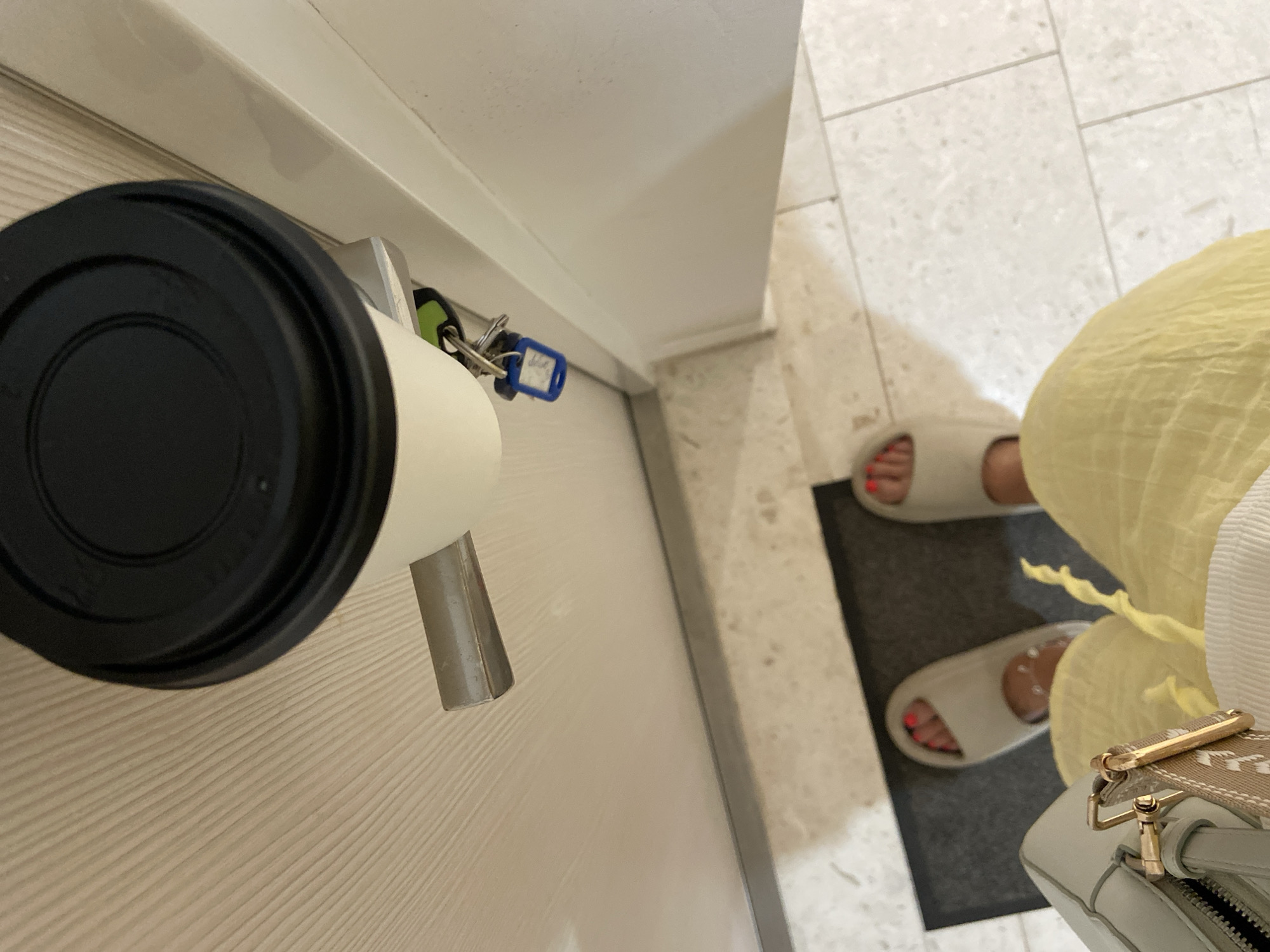 The image depicts an interior setting. Visible are a white door with a black-topped coffee cup hanging from the doorknob, and keys attached. Partial view of a person wearing white sandals with red nail polish and a yellow garment is present, along with a light-colored handbag. The floor is tiled and has a dark mat. No details conclusively indicate the location in Budva, Montenegro.