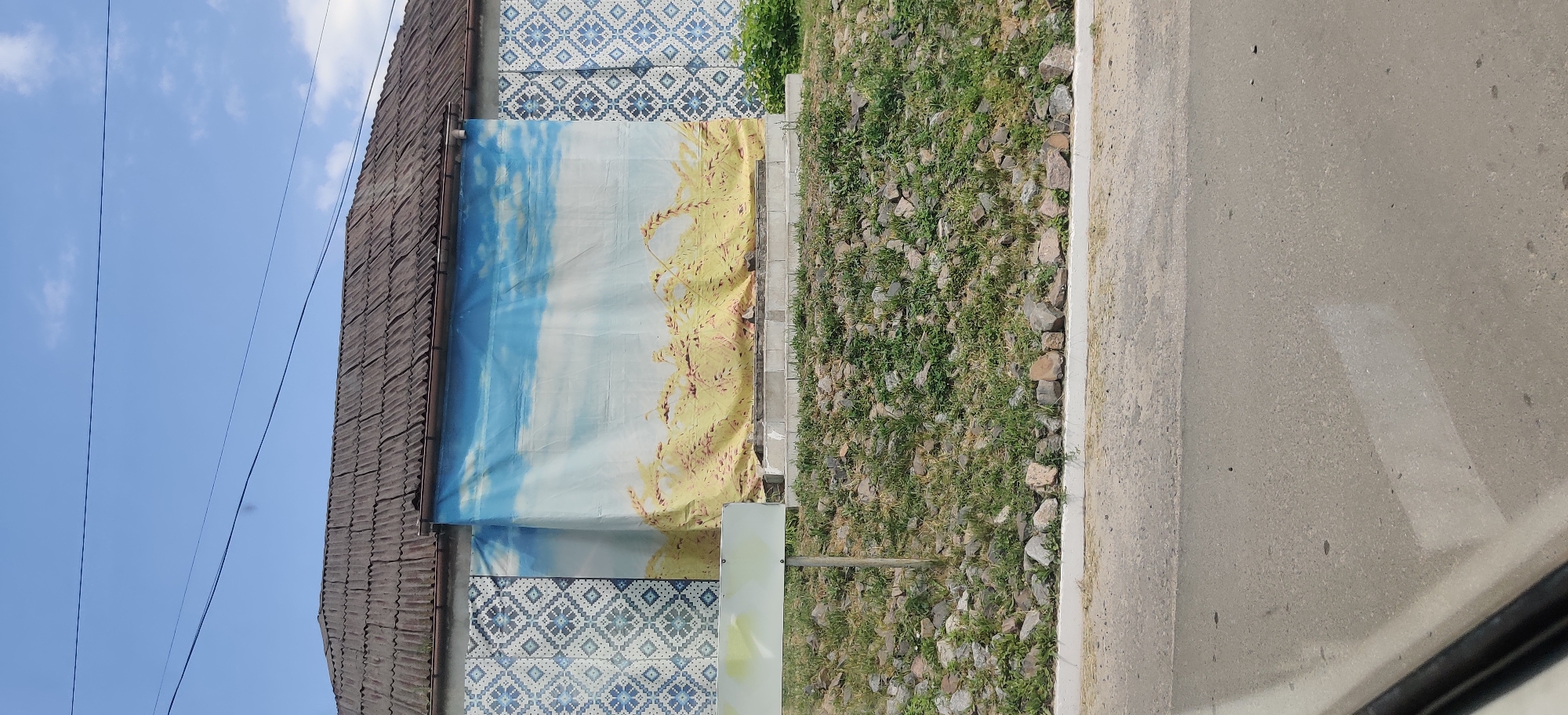 The image shows a building with a corrugated roof and a wall decorated with a blue and white patterned tile mosaic on the left side. A large banner depicting a landscape of wheat fields under a blue sky with clouds is hung across the central part of the wall. In front of the building, there is a sloped area covered with grass and rocks, with a white rectangular sign on a pole to the left. The foreground shows a paved road. The scene is located in Kolomak, Ukraine.