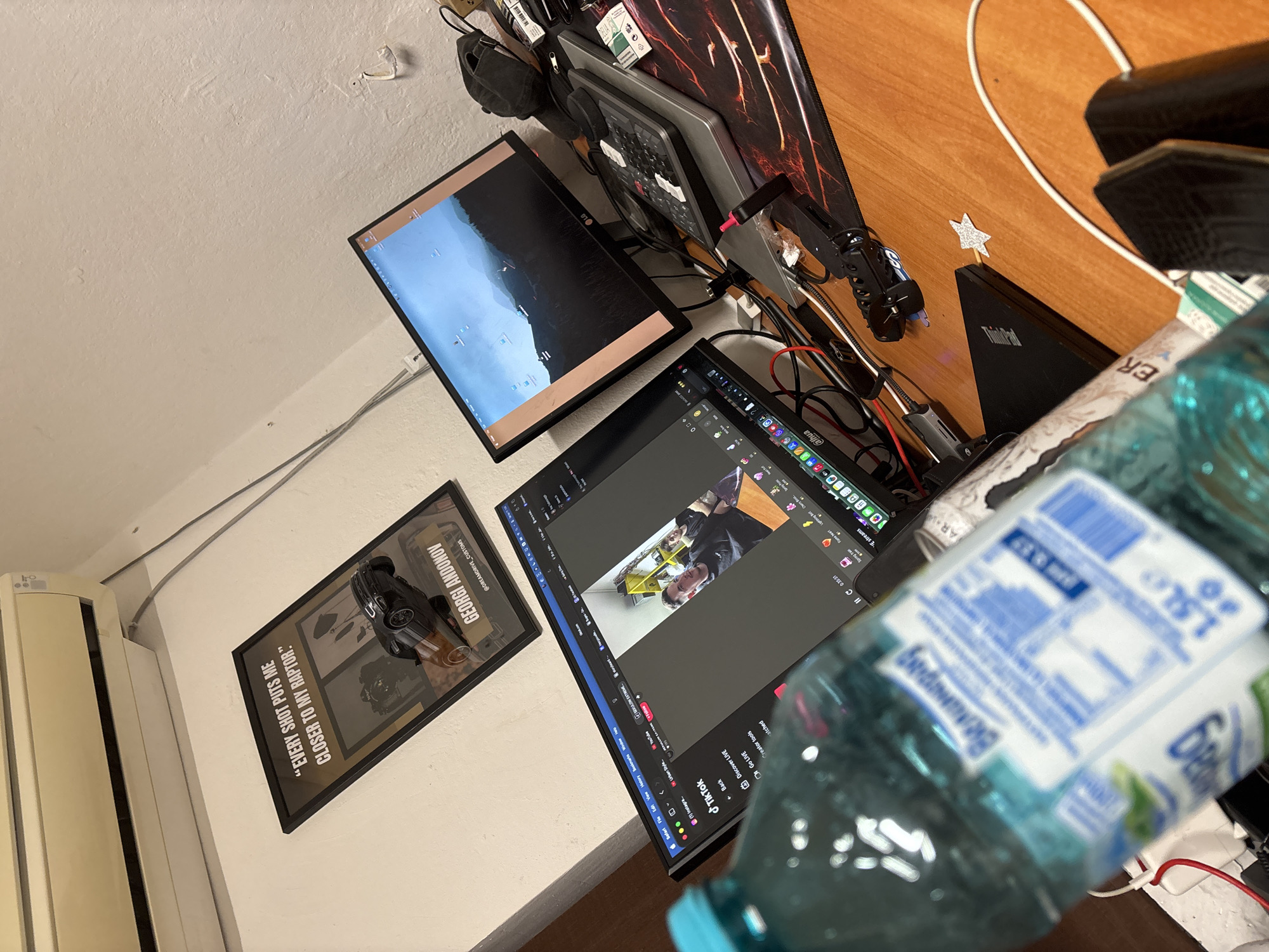 The image shows a workspace with two computer monitors, a laptop, and various accessories. One monitor displays a TikTok editing interface, while the other shows a landscape scene. A framed poster with the quote "EVERY SHOT PUTS ME CLOSER TO MY RAPTOR" and the name "GEORGI ANDONOV @DREAMDRIVE_CUSTOMS" is visible on the wall. A bottle of "Велинград" water is prominently placed in the foreground. The location is not evident based on the visual information provided.