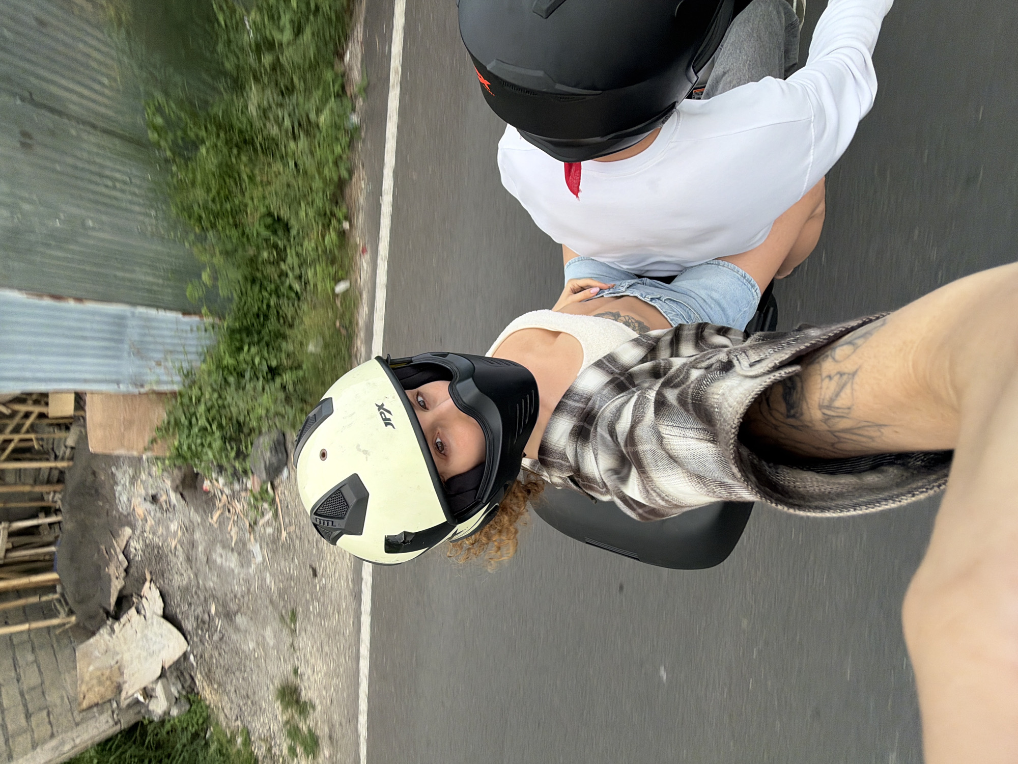 A first-person perspective selfie shows two individuals riding on a motorcycle on a paved road. The person in the foreground is capturing the image with their arm extended, wearing a plaid shirt and displaying tattoos on their arm. Their left leg is visible, with a knee bent. The person in the background, seated in front, is wearing a light-colored helmet with a black face mask, a white tank top, and denim shorts. Their curly, light brown hair is visible. They are looking back at the camera. Behind them, another person wearing a black helmet is seated, with their head and torso visible, wearing a white long-sleeved shirt. The motorcycle appears to be moving.
The environment includes a road with a white line marking the edge, bordered by lush green vegetation on the right side. On the left side of the road, there is a construction site with exposed concrete, piles of dirt and sand, wooden beams, and corrugated metal sheeting. The overall scene suggests an outdoor setting possibly in a developing area within North Kuta, Indonesia, indicated by the construction elements and tropical foliage. The lighting is overcast, contributing to a slightly muted color palette.