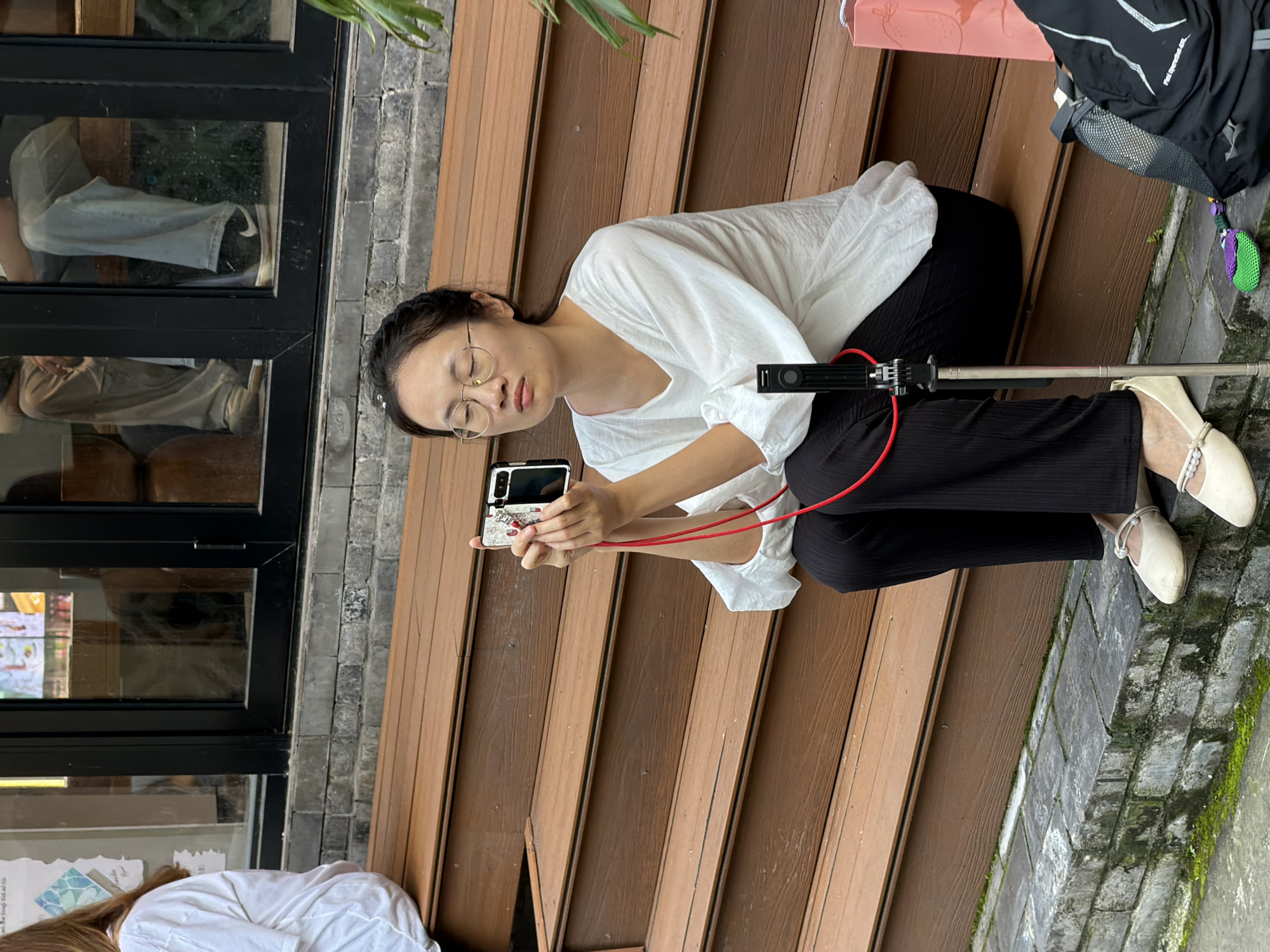 A woman sits on wooden steps holding a smartphone and a selfie stick, with a red strap around her neck, in Yuzhong District, China. She is wearing a white, loose-fitting shirt, black pants, and light-colored flat shoes. A black backpack and a pink paper bag are on the ground beside her. The background shows a building with dark window frames and reflections of people inside. The steps are made of brown wood planks, and the ground in front is paved with grey bricks with patches of green moss.
