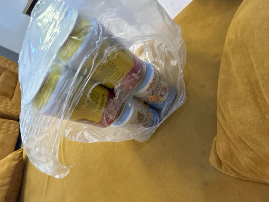 In an interior setting in Egbe, Nigeria, a clear plastic bag containing multiple beverage containers rests on a yellow-gold upholstered surface. The bag holds two primary types of containers: an upper layer of at least four cylindrical containers with yellow labels and white lids, and a lower layer of at least two light blue cylindrical containers. One of the lower containers clearly displays "Chivita Active ZEST" on its label. The upholstered surface exhibits a textured fabric and includes a visible cushion in the foreground, indicating a sofa or armchair. No individuals are visible.