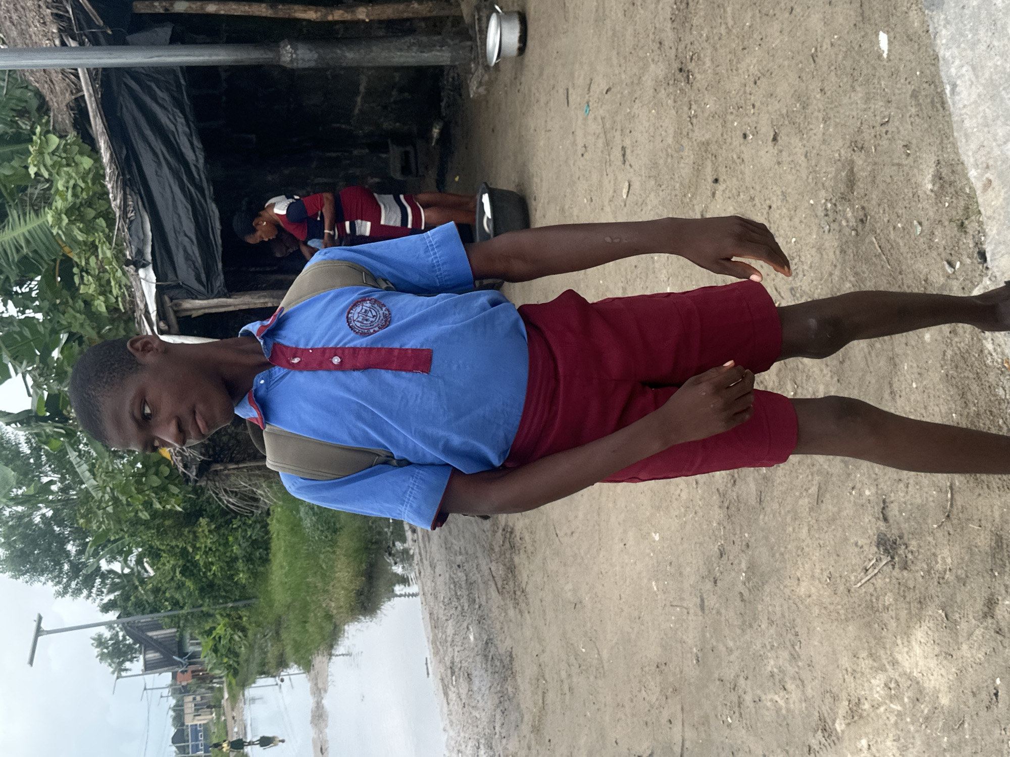 A young boy in a blue and red school uniform stands in a dirt path on an overcast day. He wears a light brown backpack. To the left of the boy, a flooded dirt road with some vegetation and buildings is visible. In the background to the right, a woman in a striped dress stands near a simple structure with a dark tarp roof. Two metal pots are placed on the ground near the structure. The scene appears to be in a neighborhood in Badagry, Nigeria.