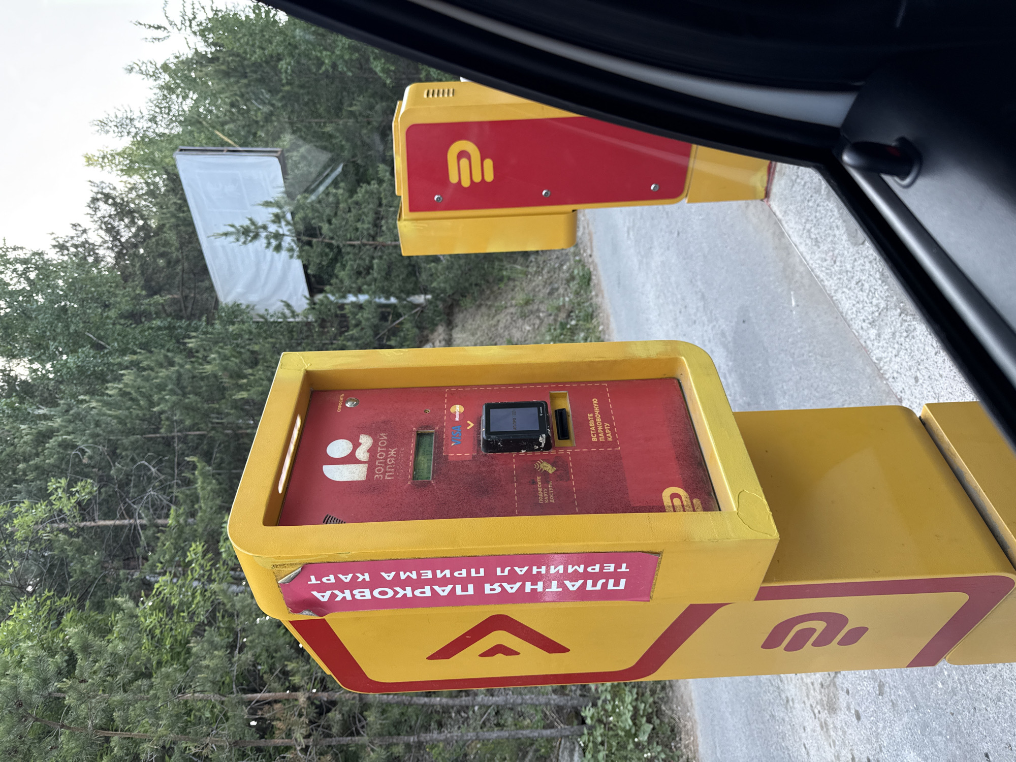 The image shows a payment terminal for parking, likely located at the entrance or exit of a parking area. The machine is predominantly yellow with red accents and features a prominent logo. The terminal accepts card payments as indicated by the inscription "ПЛАТНАЯ ПАРКОВКА ТЕРМИНАЛ ПРИЕМА КАРТ" (Paid Parking Card Acceptance Terminal). There is also a card reader and slots for inserting parking cards. Additional text in Russian is visible, including "ВСТАВЬТЕ ПАРКОВОЧНУЮ КАРТУ" (Insert Parking Card), "ПОДНЕСИТЕ КАРТУ ДОСТУПА" (Present Access Card), "СПРОСИТЬ" (Ask), and "Золотой пляж" (Golden Beach). The terminal is located near trees and foliage, with a white structure visible in the background through the foliage. The scene is viewed from inside a car. Dom Otdykha Zolotoy Plyazh, Russia is the location in the city.