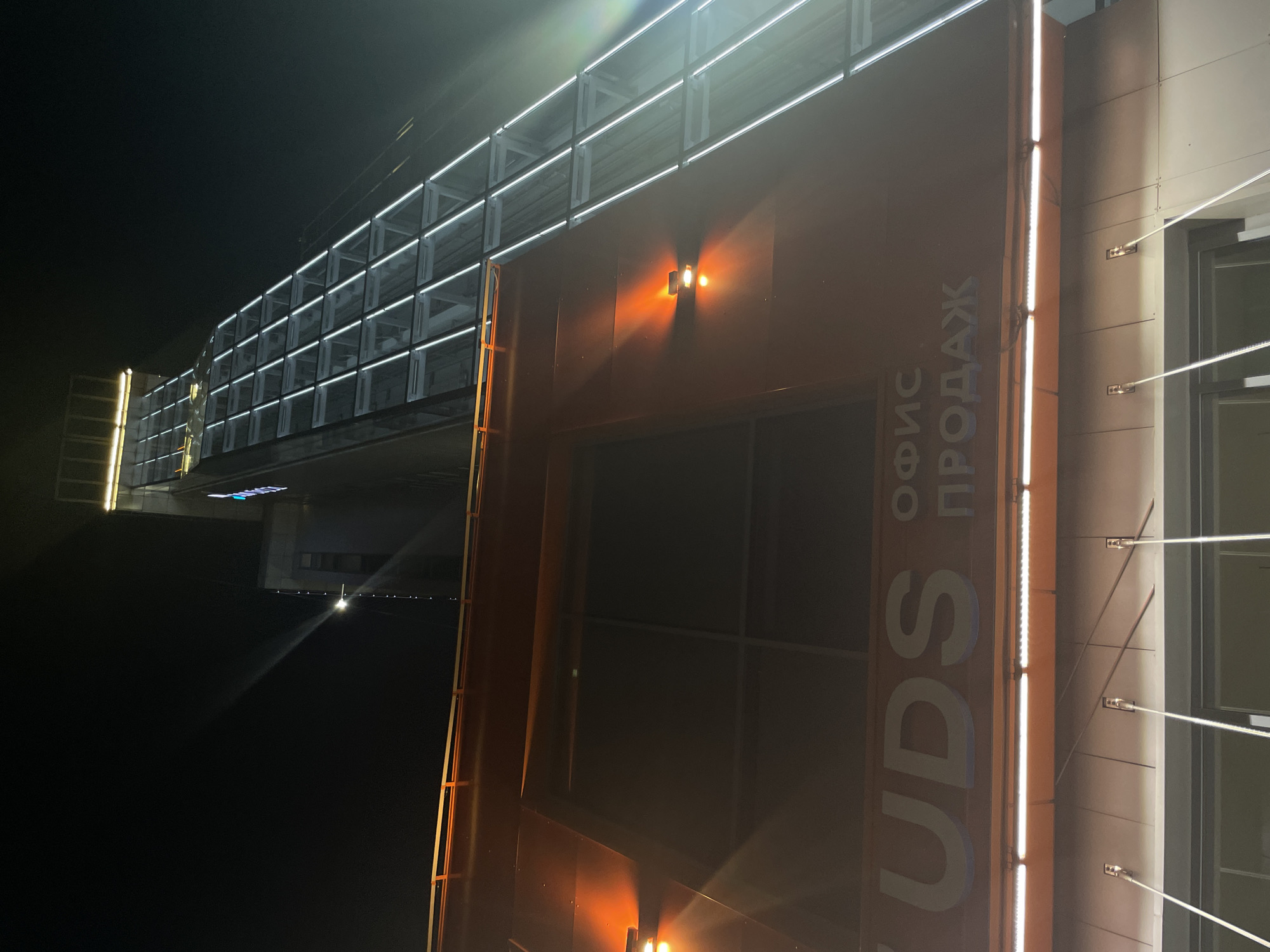 This is a photograph of a modern building facade at night. The building is illuminated by numerous white LED lights, some of which are integrated into the structure and others that are mounted vertically. There are also two warm-toned lamps on the facade, casting an orange glow. The building appears to be tall and made of metal and glass. A long walkway or bridge, also lined with white lights, extends from the building into the darkness. The sky is black, indicating nighttime. There is a prominent sign on the side of the building with Russian text that reads "UDS ОФИС ПРОДАЖ", which translates to "UDS OFFICE SALES". The overall impression is one of a sleek, contemporary commercial or office space, brightly lit against the night sky. The lighting suggests it is either a functional space or designed for aesthetic appeal.