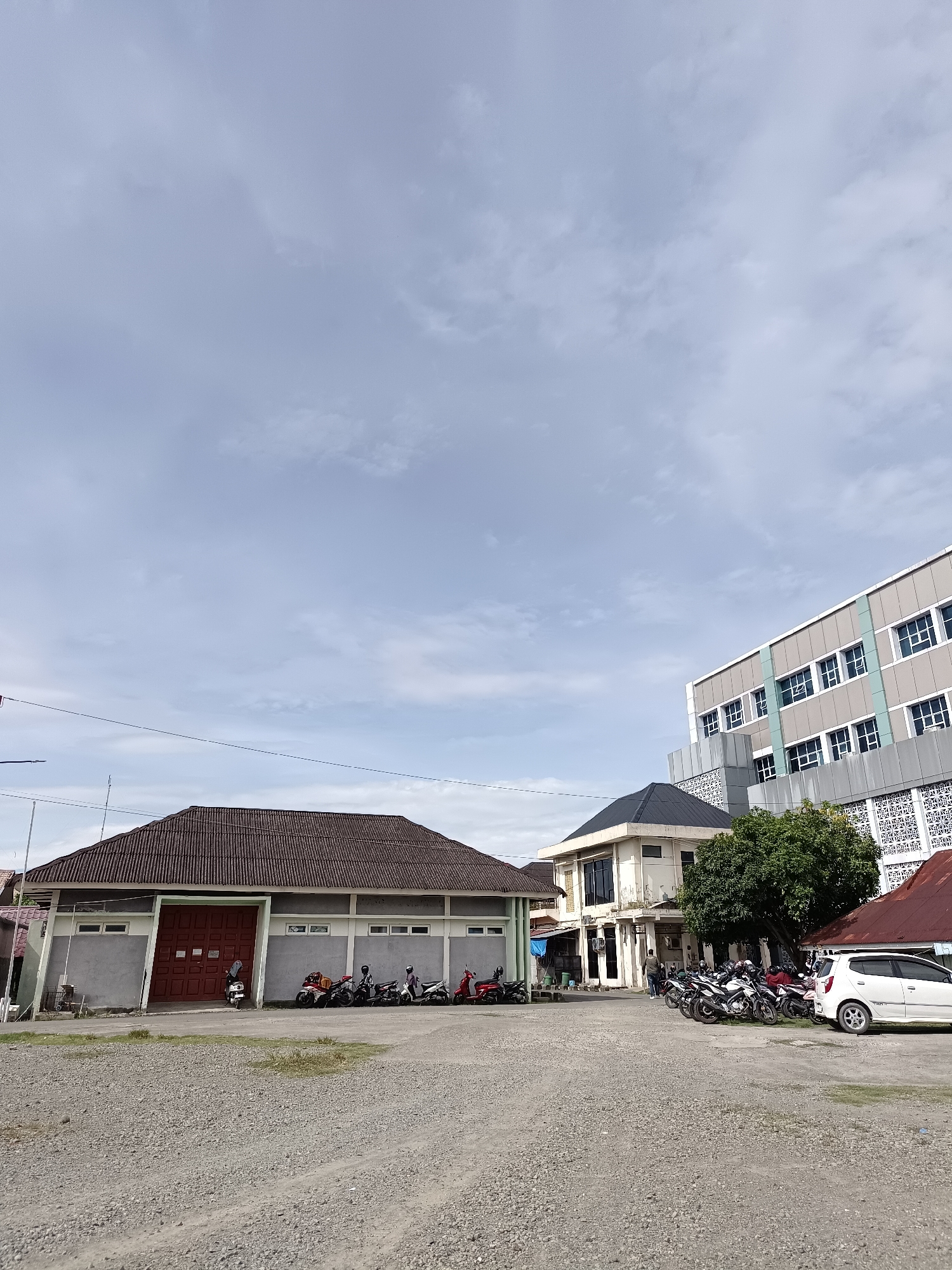 The image depicts an outdoor scene with several buildings and parked vehicles under a cloudy sky. In the foreground, a gravel lot is visible. To the left, a long, single-story building with a dark, corrugated roof and a large red door dominates the frame. Several motorcycles are parked in front of this building. Further to the right, a multi-story building with a modern facade, featuring numerous windows and a light gray and teal color scheme, rises high into the sky. Between these two structures, a smaller, lighter-colored building with a dark roof is situated. A white car is parked to the right of the motorcycles, near the base of the multi-story building. Trees and other foliage are present, particularly around the smaller building. A few individuals can be seen near the vehicles. The overall environment appears to be an institutional or commercial area within the city of Mesjid Reuntoh, Indonesia.