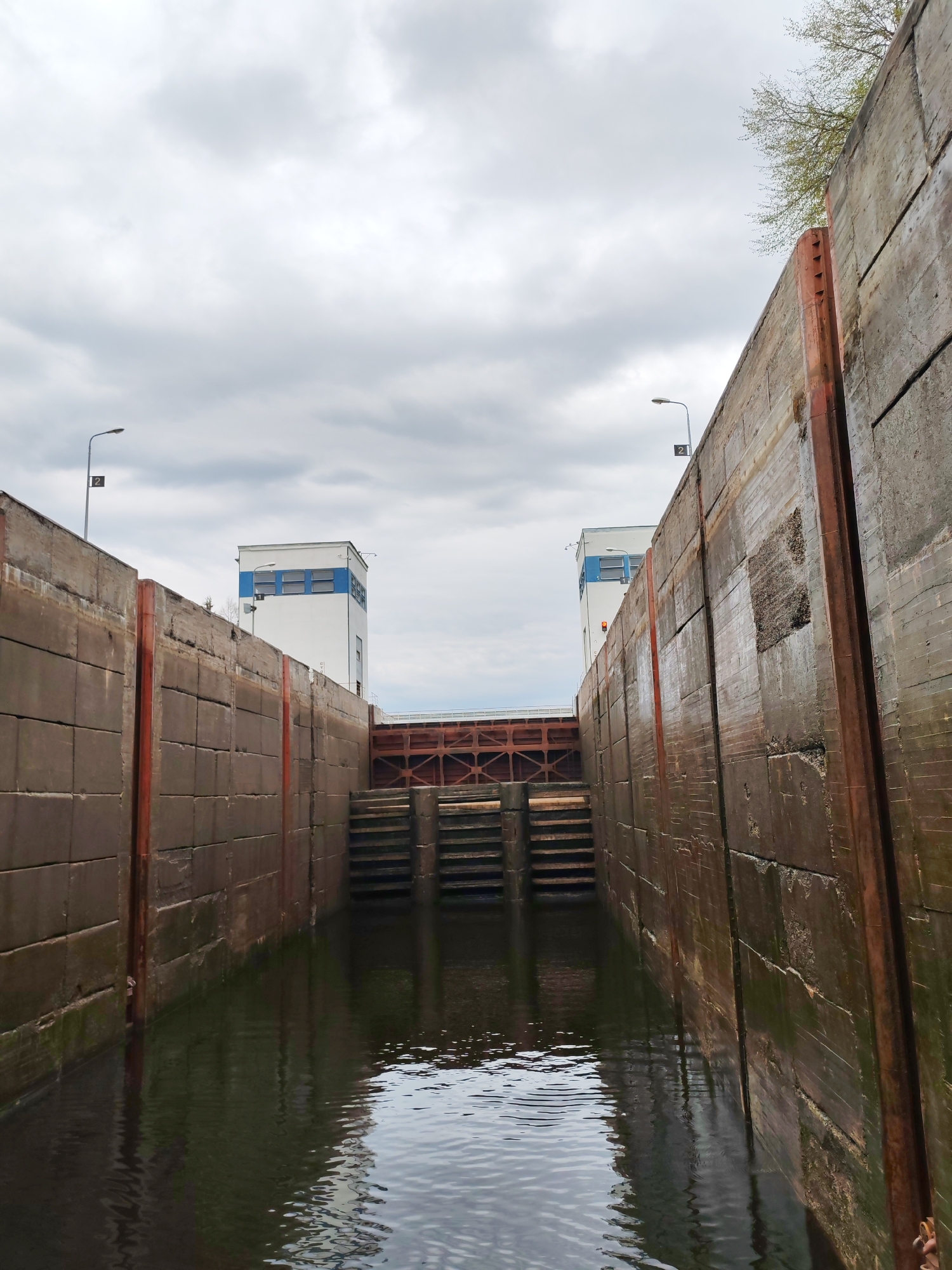 A low-angle, eye-level shot shows a water passage or lock at an unknown location. Two concrete walls rise from the water, with metallic red vertical supports. Across the passage, concrete steps are visible beneath a metal structure, with two buildings beyond. Overhead, a cloudy sky is visible. Two light posts flank the passage entrance. The water's surface reflects the scene.