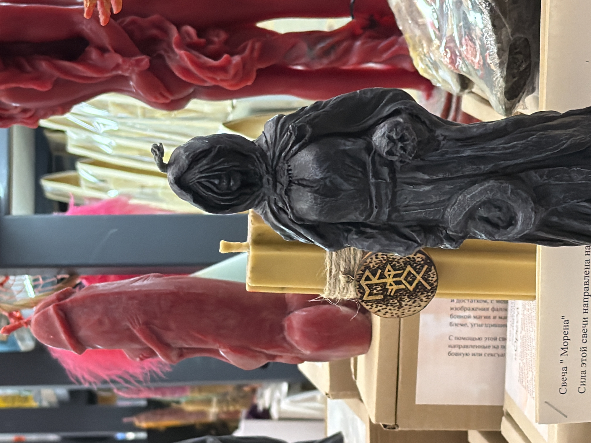 This image depicts a shelf displaying various candles, possibly in a shop or market in Moscow, Russia, based on the Cyrillic text on the label. The candles include a black figure resembling a cloaked character, a red figure resembling a reclining person with exaggerated features, and other red and yellow candles in the background. A tag with a geometric design is attached to one of the candles. The scene appears to be indoors, with a focus on the merchandise.
