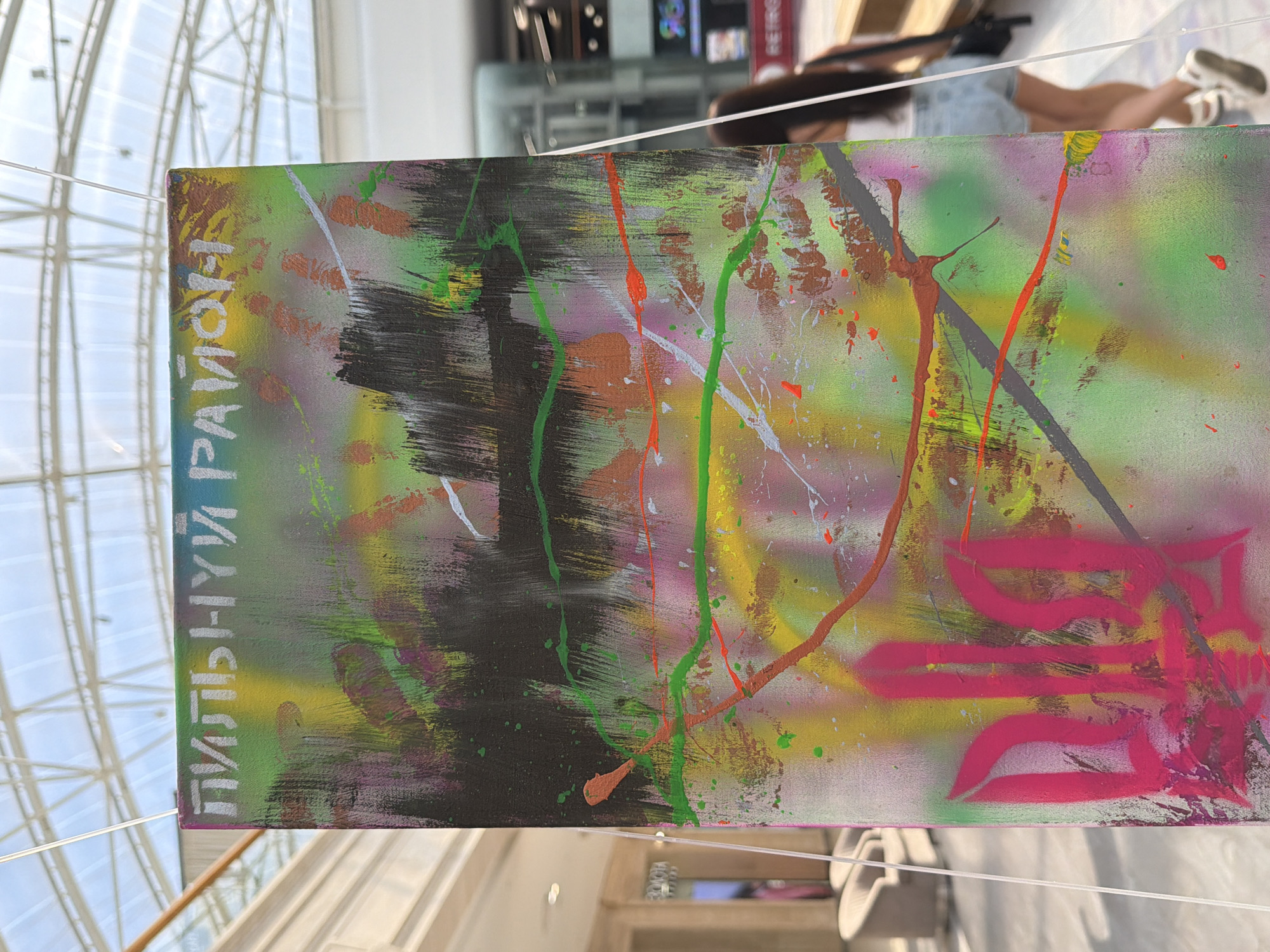 A close-up, vertical, eye-level shot features a vibrant, abstract painting displayed indoors, possibly in a shopping mall with a glass ceiling visible in the background. The painting is composed of energetic strokes and splatters of various colors, including pink, green, yellow, black, and orange. A prominent pink trident symbol, the national coat of arms of Ukraine, is spray-painted in the lower-left quadrant of the artwork. White text, appearing to be in Cyrillic, is visible at the top of the painting. The artwork is suspended by thin wires. In the blurred background, a woman wearing a white top and denim shorts walks away from the camera. The environment suggests a public space with architectural elements like railings and distant shops.