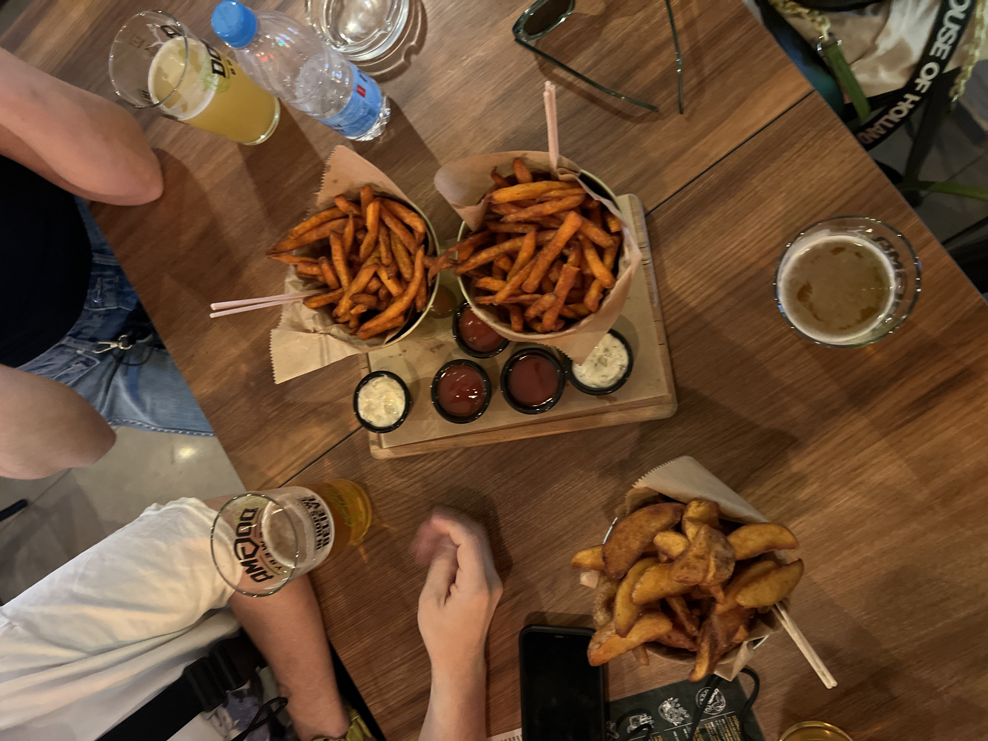 The image depicts a table at a restaurant, likely located in Belgrade, Serbia. The table is made of dark wood, and there are several food and beverage items on it. Two people are seated at the table, one with their legs visible on the left side of the frame, and the other with their arm and hands visible on the bottom of the frame. The table is cluttered with two servings of sweet potato fries served in paper cones inside metal containers, a serving of potato wedges, a wooden tray with three sauces, two glasses of beer, a bottle of water, a pair of sunglasses and a mobile phone. The scene is a casual dining setting.