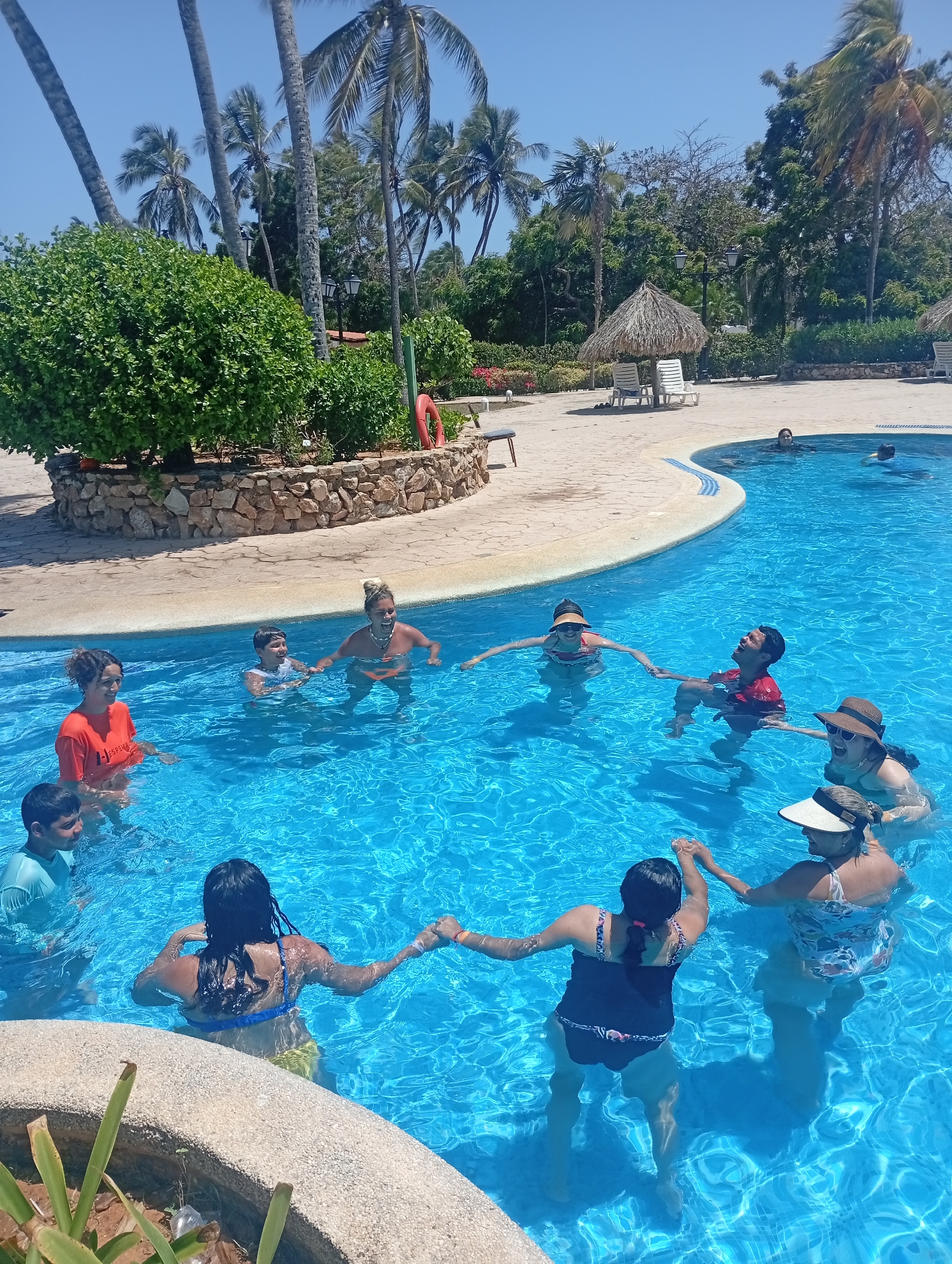 A group of people, including adults and children, are in a swimming pool, holding hands in a circle. The pool is outdoors and surrounded by trees, including palm trees, and a thatched-roof structure. Some people are wearing swimwear and hats. Two other people are swimming in the pool farther away from the group. The location appears to be a resort, though the city and country are unknown.