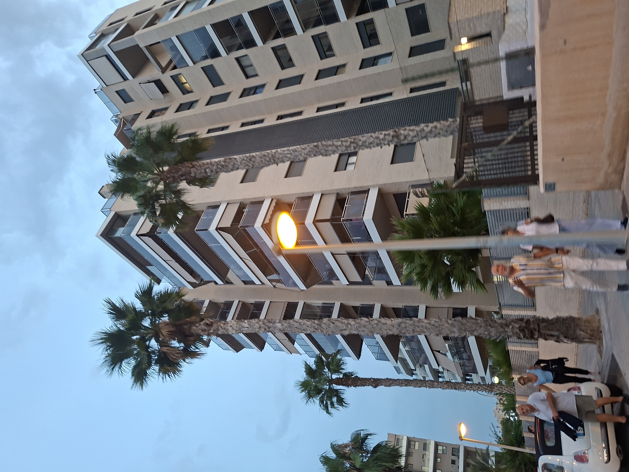 In Alicante, Spain, a street scene unfolds under a cloudy sky. Palm trees punctuate the foreground, with their fronds swaying gently. A modern apartment building, clad in beige brick and featuring numerous balconies with glass railings and retractable awnings, dominates the background. A streetlamp with a bright yellow light illuminates a portion of the sidewalk.
Several people are present. On the left, a man wearing a light grey polo shirt and khaki shorts stands near a white Fiat 500 car. He is holding a dark jacket. To his right, a woman in a blue sleeveless top and dark pants walks away from the car, carrying a black bag. Further to the right, a man in a striped short-sleeved shirt and light-colored pants is visible, gesturing with his right hand. Another person, partially obscured, walks behind him.
The environment suggests a residential urban area, likely in the late afternoon or early evening. The architecture of the building and the presence of palm trees are characteristic of coastal Spanish cities.