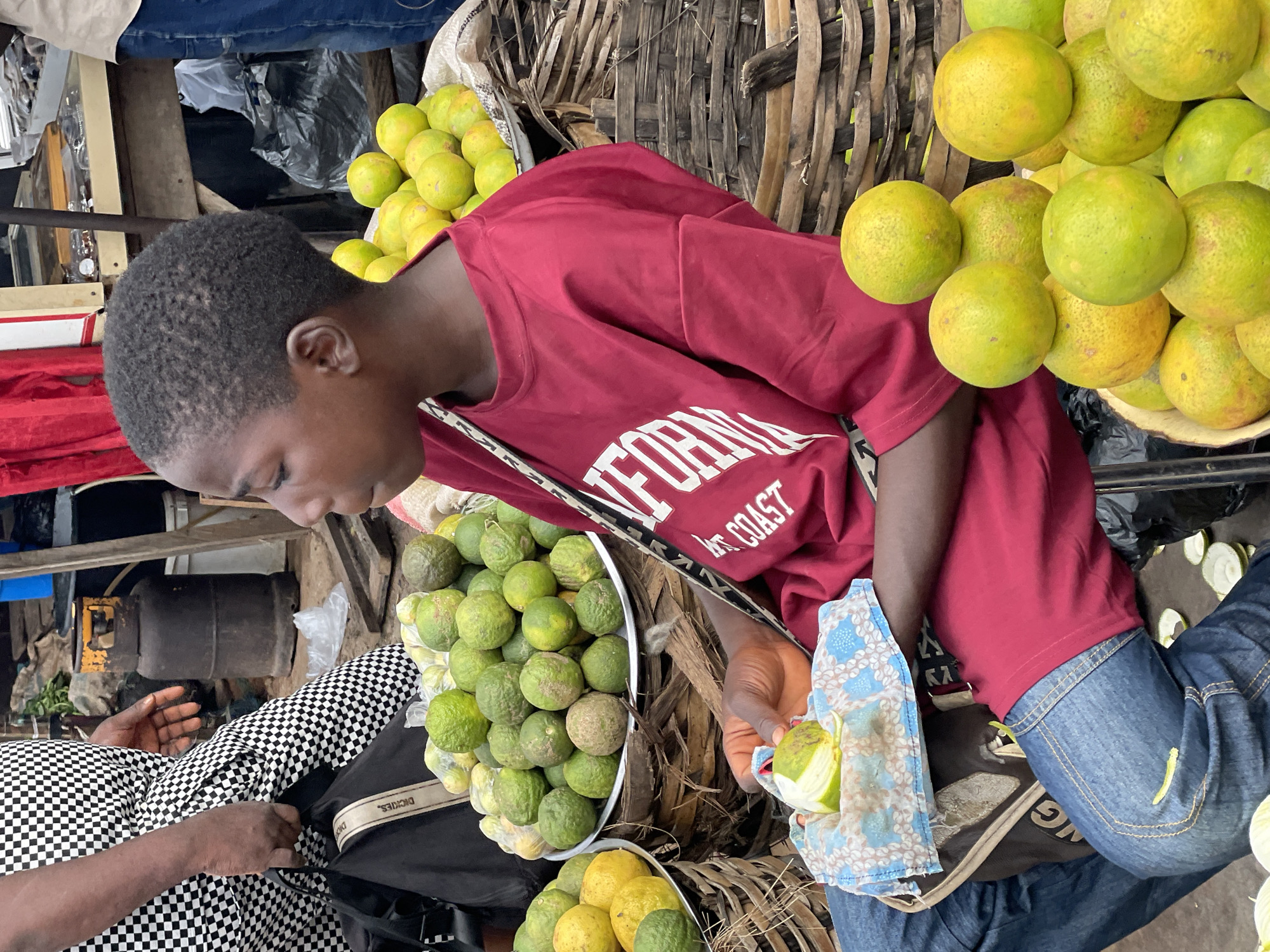 A young Black person sits in an outdoor market in Nkpor, Nigeria, surrounded by fruits and vegetables. The person wears a red t-shirt with white lettering that reads "CALIFORNIA COAST" and blue jeans. In their right hand, they are peeling a green fruit, possibly a lime or a small orange, which rests on a blue and white patterned cloth.
The market scene features several large woven baskets filled with green and yellow fruits. In the foreground, a basket overflows with bright yellow-orange fruits, some stacked in pyramids. Behind this, a large shallow metal bowl is filled with green, round fruits, likely limes or small oranges, with some scattered on the ground. To the left, another basket holds yellow fruits.
In the background, an individual wearing a black and white checkered garment is visible, holding a black bag. A yellow gas cylinder stands near a wooden structure. The ground appears to be dirt or compacted earth, with various items scattered around, including discarded fruit peels. The overall environment suggests a bustling, open-air market with fresh produce for sale.