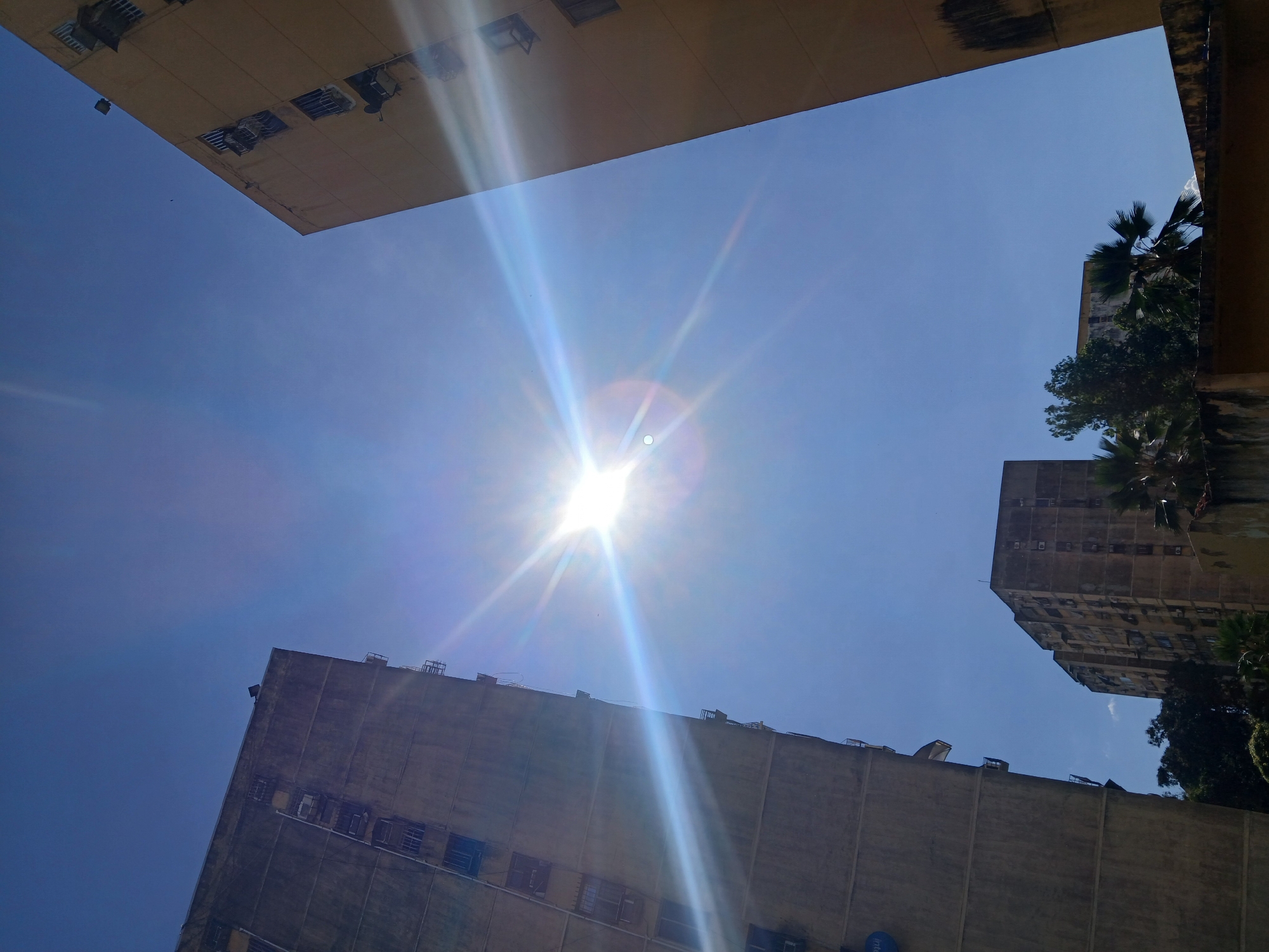 The image depicts an outdoor urban scene in Puerto Ordaz, Venezuela, characterized by a clear, bright blue sky and harsh sunlight. Two tall, weathered apartment buildings dominate the foreground, angled towards the viewer from the left and right sides of the frame, respectively. The building on the left appears to be concrete-gray, with numerous visible air conditioning units and balconies. The building on the right is a pale yellow, also showing signs of age, with similar architectural features. The sun is positioned prominently in the upper center of the image, causing lens flare and bright streaks of light that extend across the sky and the buildings. In the distance, a third, slightly shorter apartment building can be seen, along with lush green trees and palm fronds, suggesting a tropical or subtropical environment. The overall impression is one of a sunny day in a densely populated urban area.