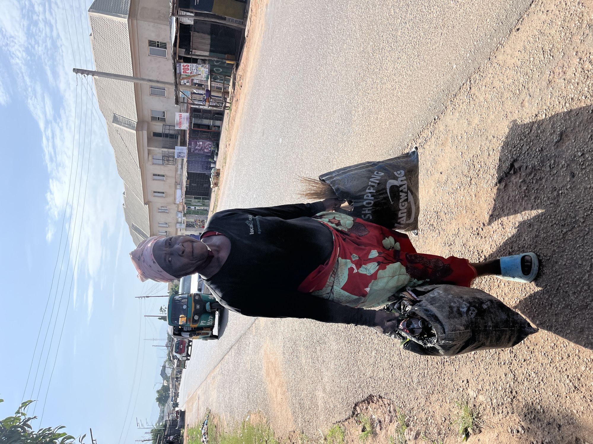 A mature Black woman in Jos, Nigeria, stands on the roadside, holding two large black trash bags. She wears a dark long-sleeved shirt, a red and green patterned skirt, a light pink headscarf, and blue sandals. One bag contains a broom. Behind her, a road stretches into the distance with buildings, power lines, and vehicles (including a "Keke" tricycle taxi) visible. The sky is partly cloudy. The ground is a mix of pavement and dirt.