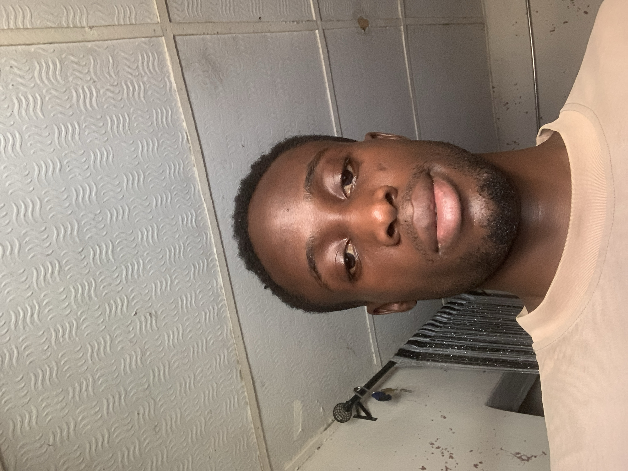 The image captures a close-up portrait of a young man of African descent, whose head is tilted almost 90 degrees to the left. He looks directly at the camera with a neutral expression, his lips parted slightly as if on the verge of a smile. He has short, dark hair and a short beard or stubble. He is wearing a light-colored, possibly beige or cream-colored, t-shirt with a visible ribbed collar.
The background is dominated by light-toned, textured panels that resemble acoustic ceiling tiles or decorative wall panels. These panels feature a uniform, repeating pattern of small, wavy lines arranged in rows. Visible seams delineate these panels. In the lower right section of the image, beneath the man's chin and neck, there is a dark, somewhat reflective surface, likely a countertop or table. This surface is speckled with numerous small, dark particles, suggesting dirt or debris. A small, dark object, possibly part of a faucet or plumbing fixture, with a circular gridded top and a handle-like structure, rests on this speckled surface. Behind the man's right shoulder (on the left side of the image due to the tilt), a darker, possibly ribbed or corrugated surface is partially visible. The overall setting appears to be an indoor environment, possibly a utility space or bathroom. No text is visible in the image.