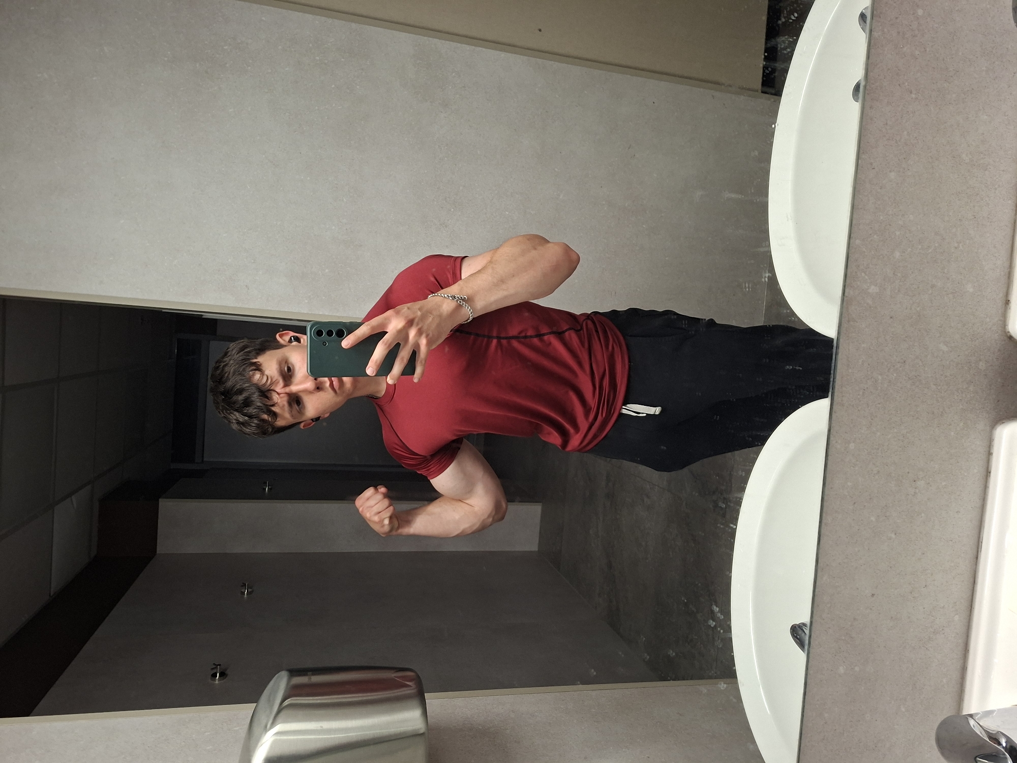 A young man takes a mirror selfie in what appears to be a public restroom or changing room in Plovdiv, Bulgaria. He is seen from the chest up, flexing his right bicep towards the camera. He has dark, curly hair and is wearing a red short-sleeved t-shirt and black athletic pants with a white detail. A dark-colored smartphone with a triple camera setup is held in his left hand, and a silver-colored chain bracelet adorns his right wrist. A dark earbud is visible in his left ear.
The setting is characterized by light gray, speckled walls and countertop, with darker gray or black tiled sections visible in the background reflection. On the right side of the image, two white ceramic sinks are partially visible, set into the countertop. In the lower-left corner of the reflection, a silver, metallic object, likely a hand dryer or soap dispenser, can be seen. The overall atmosphere is utilitarian and indoors. The man's expression is focused as he poses for the picture. There is no visible text in the image.