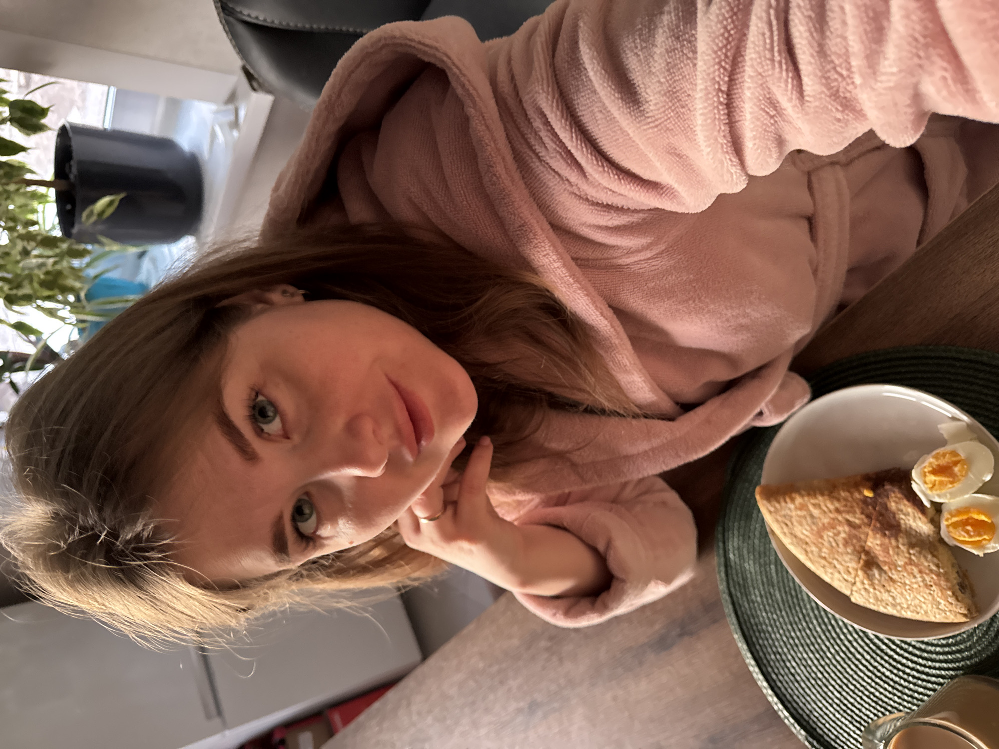 A woman in a pink bathrobe is pictured from a high angle. The location is Ферма, Russia. She is sitting at a table with a plate of food containing what appears to be a pastry item and two halves of a hard-boiled egg. A plant in a dark-colored pot is visible in the background near a window.