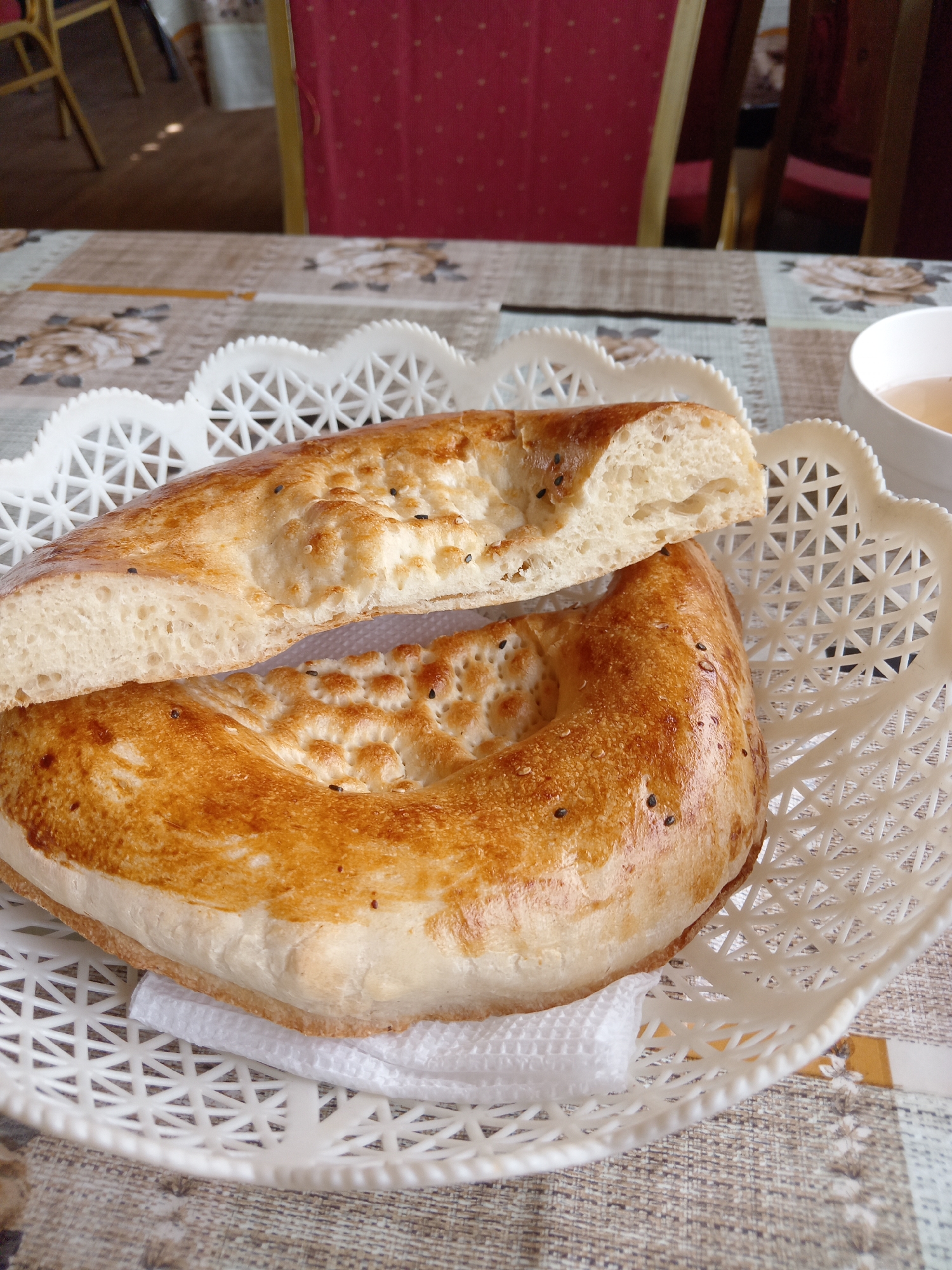 A close-up, eye-level shot focuses on a white, intricately patterned plastic basket holding two pieces of golden-brown flatbread, likely lavash or tandyr nan, sprinkled with black sesame seeds. One piece is broken in half, revealing a fluffy, airy interior. The bread rests on a white paper napkin. The basket sits on a tablecloth with a muted floral pattern in shades of beige and brown, possibly in a cafe or restaurant in the city of Ması, Kyrgyzstan. In the upper right corner, the rim of a white teacup containing a light amber liquid is visible, suggesting a beverage accompanying the bread. In the blurred background, red upholstered chairs with metallic legs are partially visible, indicating a seating area. The lighting is natural and diffused, highlighting the texture and golden hue of the bread. No people are visible in the frame.