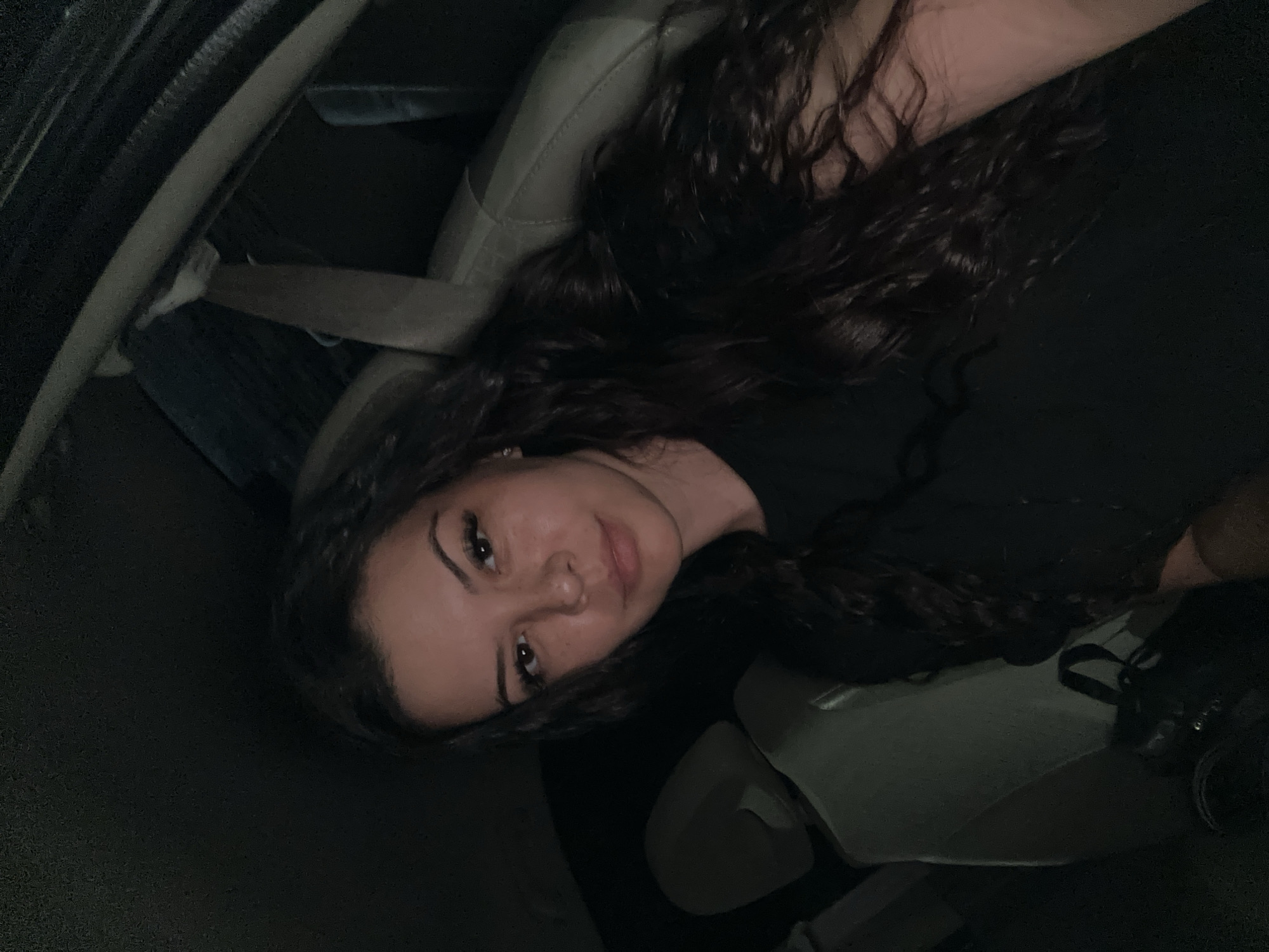 A young woman with long, dark, curly hair is pictured from the chest up, sitting in the passenger seat of a car at night. She is wearing a black t-shirt and looking towards the camera with a slight smile. The background is dark, with the headrest of the driver's seat visible to her left and the car's interior dimly lit. The image does not provide enough information to determine the location within Sliven, Bulgaria.