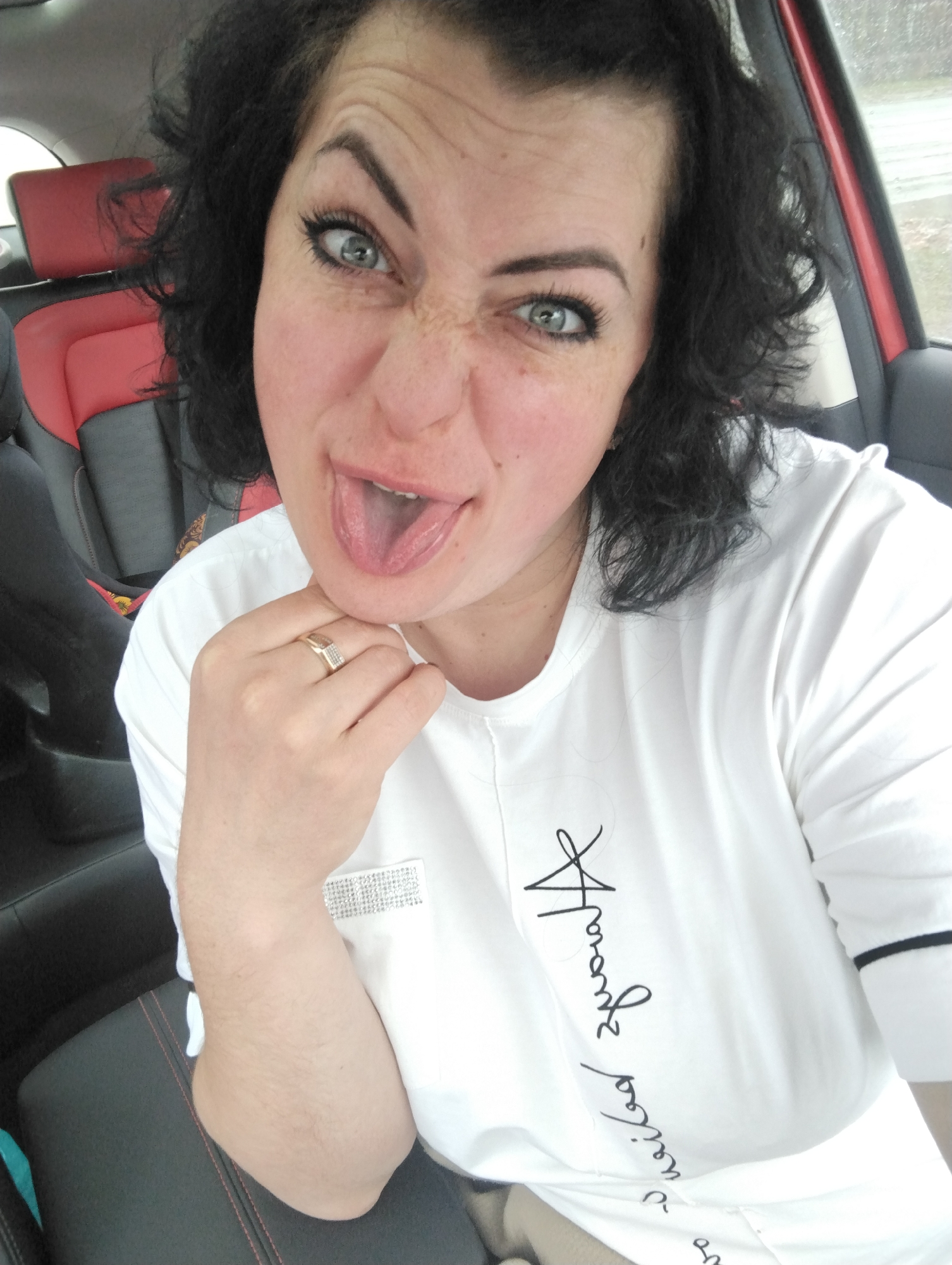 A woman with dark, curly hair and striking light-colored eyes is captured in a playful selfie inside a car, likely in Izhevsk, Russia. She is making a humorous face, sticking out her tongue and scrunching her nose, creating a cheerful and expressive mood.
She is wearing a white long-sleeved t-shirt featuring black cursive writing, part of which appears to be "Hravabe papem," and a rectangular patch of silver, sparkly embellishments on the left chest area. A gold-toned ring adorned with small clear stones is visible on her right hand, which rests under her chin.
The setting is the interior of a car, characterized by red and black upholstered seats. A child's car seat, predominantly black with red trim, is visible in the background on the left. The lighting is soft and diffused, suggesting daytime, possibly overcast weather, with a blurred view of a gray outdoor environment visible through the passenger window.