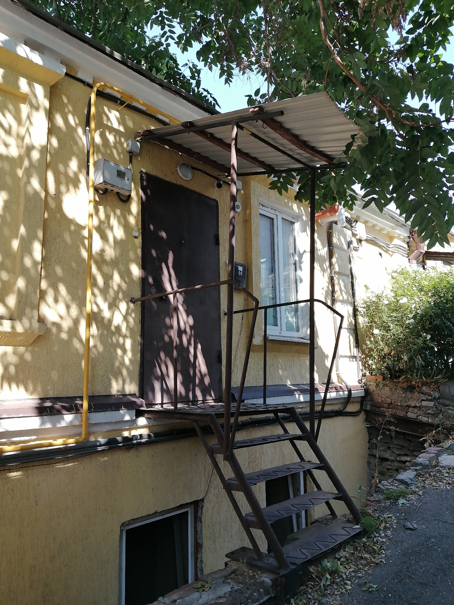 This is a medium shot, taken at eye level, of the exterior of a yellow stucco building, likely in Rostov-on-Don, Russia. The building has a dark brown door and a window with white trim to the right of the door. An aged, dark metal staircase with four steps leads up to a small, weathered metal canopy above the door. A yellow pipe runs vertically up the side of the building, with a grey electrical box attached near the top. Shadows from the tree canopy overhead are cast across the building's facade and the staircase. The ground in front of the building is paved and littered with fallen leaves and debris. To the right of the staircase, there is a lower entrance to the building, and beyond that, a brick retaining wall and dense green foliage. The sky is visible through the branches of the tree.