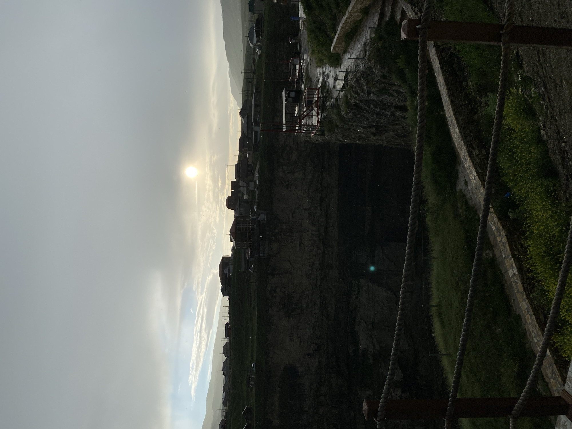 The image depicts a view overlooking a deep gorge or canyon in Khunzakh, Russia. The sun is partially obscured by clouds, casting a soft, diffused light over the scene. On the far side of the gorge, a village or town is visible with houses situated on a grassy plateau. A road or path winds along the edge of the gorge, with some structures and what appear to be viewing platforms. The foreground features a rope barrier, suggesting a safety measure for visitors. The gorge itself is dark and appears to have water at its base, though it is mostly obscured by shadow. The landscape is characterized by rugged terrain and vegetation.