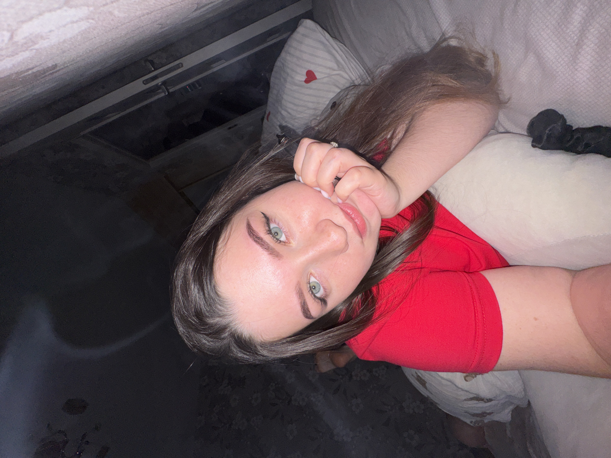 The image captures a young woman lying down in an indoor setting, likely a bedroom or living room. She has long, dark brown hair and striking light-colored eyes (appearing green or blue-green) with distinct dark eyeliner. She wears a red short-sleeved top. Her right hand, adorned with a ring on her ring finger, is casually placed near her mouth, with her fingers lightly touching her lips.
The woman is looking directly at the camera, giving a relaxed and direct gaze. She is positioned on what appears to be a bed or sofa, with her head resting on a pillow that has a pattern of red hearts on a striped background. Another light-colored, possibly white or off-white, pillow is visible near her arm. A black hair accessory, possibly a scrunchie, lies on the bedding next to her.
The lighting in the image is dim, suggesting it was taken in the evening or at night. The background on the left side of the frame shows dark elements, possibly furniture, a wall panel, or a window with a blind or shutter. The overall atmosphere is intimate and candid. No visible text is present in the image. The provided location context is Perm, Russia.