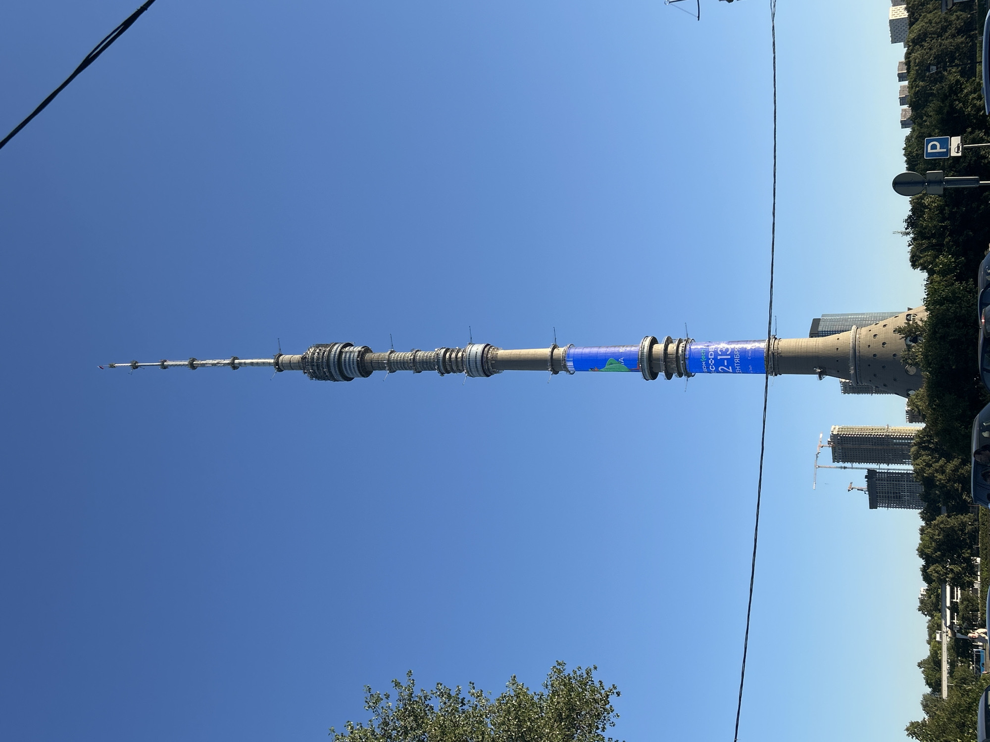 The image shows the Ostankino Tower in Moscow, Russia, on a clear day. The tower is a prominent television and radio tower. A blue digital display is visible on the tower, featuring text that appears to be in Cyrillic and English. Below the tower, a line of trees and some construction sites with cranes are visible. A few cars are parked or driving in the foreground, and a parking sign with a 'P' is also present. The sky is a bright, cloudless blue.