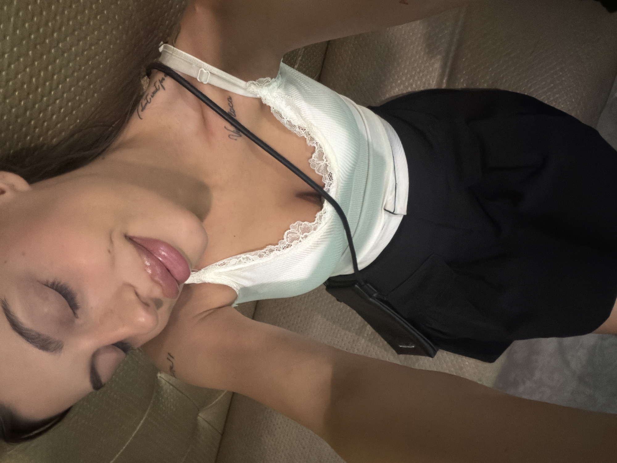 A woman with closed eyes and full lips is captured in a selfie. She is wearing a white lace-trimmed tank top and black shorts. A small black purse is slung across her body. The background appears to be a textured couch in a dimly lit indoor setting. The woman has tattoos on her left shoulder and collarbone. The image does not contain any information that would indicate a location in the city of Burgas, Bulgaria.