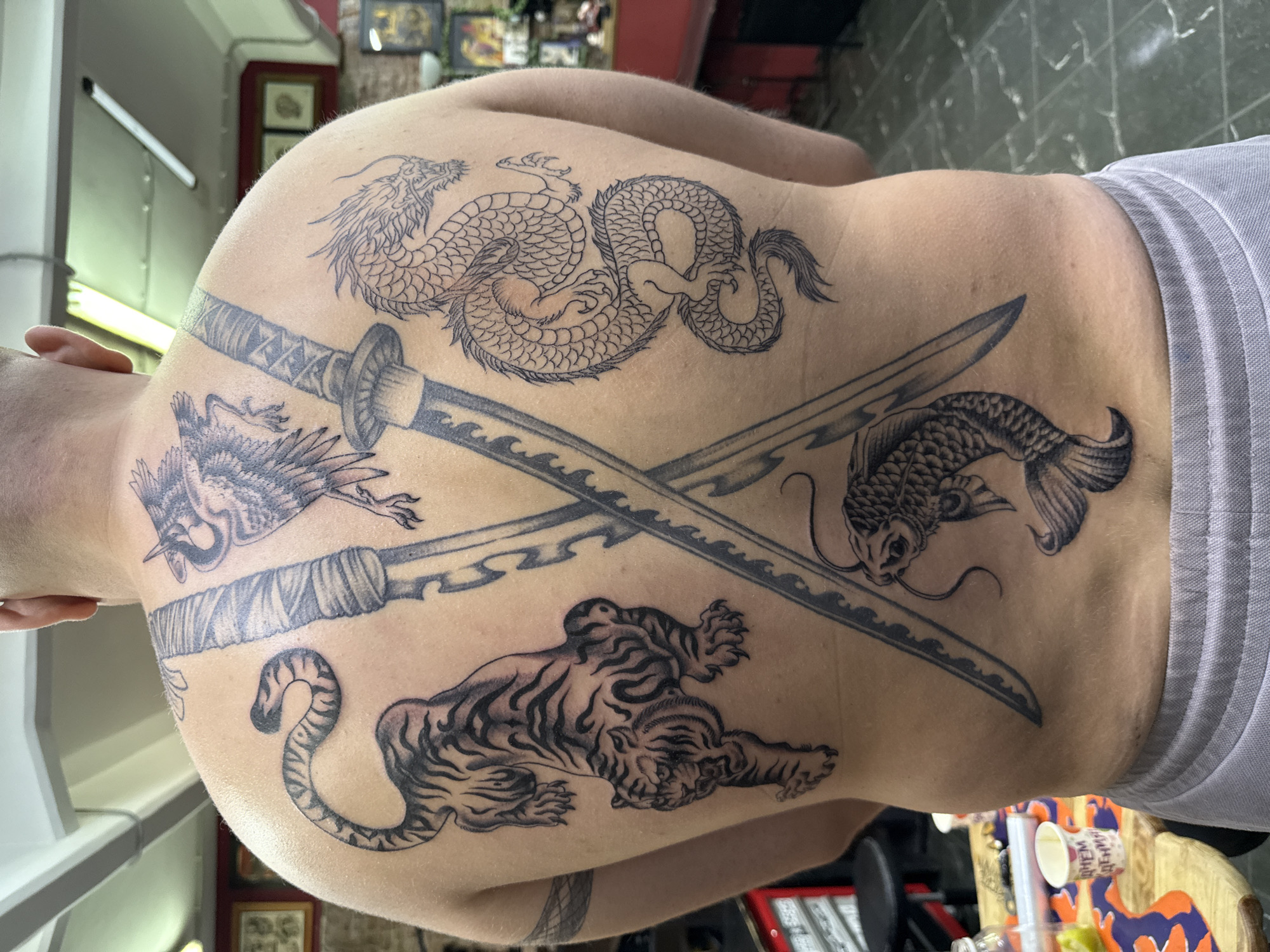 A male individual is depicted from the back, shirtless, revealing a complex array of black ink tattoos across his entire back. He wears light grey athletic shorts. The tattoos include a crouching tiger on the viewer's left, an elongated, coiled dragon on the viewer's right, and two crossed samurai swords centrally positioned between them. Above the crossed swords, an avian creature with spread wings is tattooed on the upper right shoulder area, while a koi fish swimming downwards is located below the swords on the lower back. A patterned band-style tattoo is visible on his upper left arm.
The scene is an indoor setting, consistent with a tattoo studio. The background features light-colored walls, with several dark-framed pictures visible. Bright fluorescent light fixtures are mounted on the ceiling. In the lower left foreground, a wooden table holds a clear bottle, an orange and blue patterned mat, and a white paper cup with red, partially visible Russian text reading "ДНЕМ РОЖДЕНИЯ" (Happy Birthday). A black stool is also present. The far background includes a red upholstered bench and display shelves with various items. The floor is tiled. While the Russian text indicates the location is in Russia, the specific city of Moscow cannot be confirmed from the visual evidence.