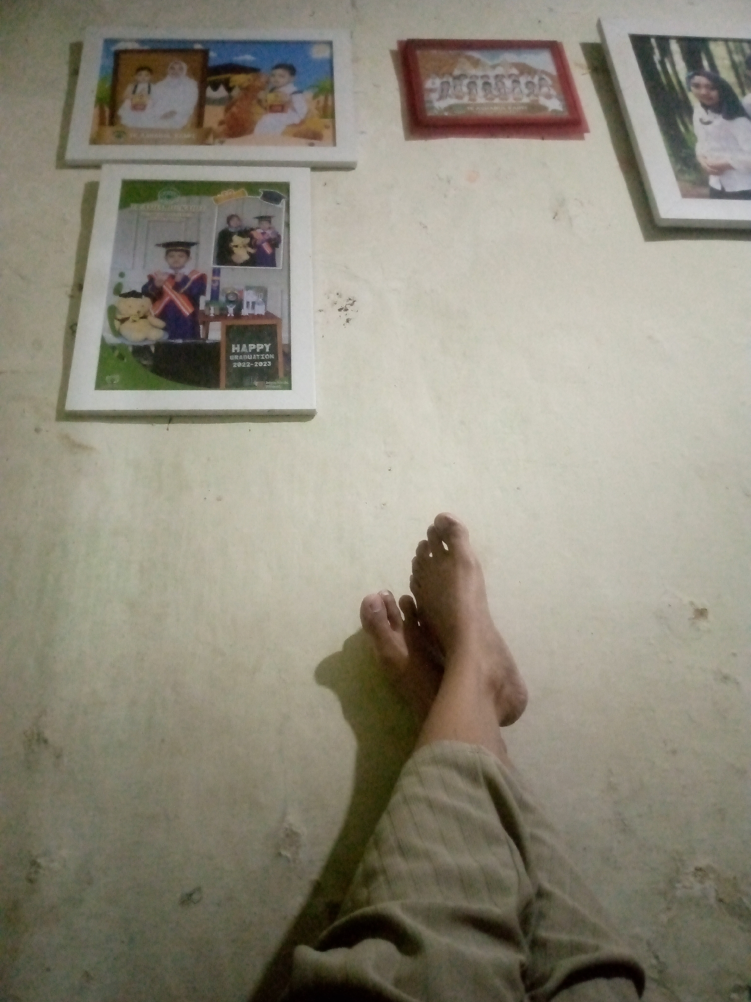 The image shows a wall with several framed pictures. In the foreground, a person's bare feet and legs, wearing beige pants, are visible, suggesting they are resting. One foot is positioned at an angle, and the other is slightly behind it. The wall itself is textured and appears to be painted a light color.
The framed pictures on the wall are:
* A horizontal frame containing two children dressed in white, with a backdrop suggesting a traditional Indonesian setting.
* A vertical frame displaying a graduation collage. It features a child in a graduation cap and gown, a teddy bear, and text that reads "HAPPY GRADUATION 2022-2023".
* A small, dark red frame containing a group photo of individuals in what appears to be a formal setting.
* A large, white frame with a portrait of a young woman wearing a white top.
The lighting in the scene casts shadows, indicating the light source is likely to the right of the person's feet. The overall impression is of a domestic indoor setting.
Based on the provided information, this scene is located in Laksanamekar, Indonesia.