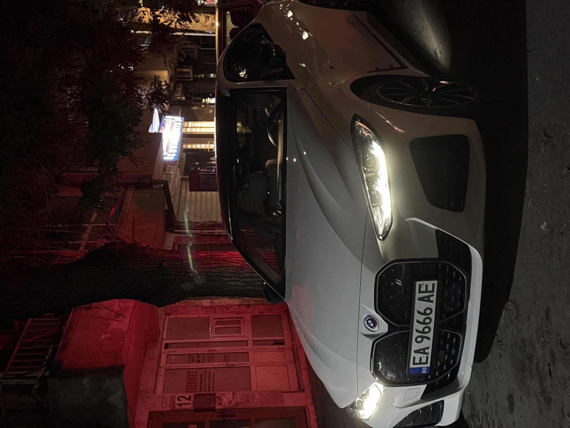 The image displays a white BMW car, likely a 4 Series, parked at night on an urban street in Plovdiv, Bulgaria. The car's bright headlights are illuminated, casting light onto the immediate foreground. The Bulgarian license plate, clearly visible on the front grille, reads "EA 9666 AE" with the "BG" country code on the blue EU strip. The BMW logo is prominent on the large kidney grille.
To the left of the vehicle, a building facade is visible, bathed in a strong red light. This illumination highlights details such as windows, doors, and a partial sign that reads "INTERNET" and the number "12". A large, dark tree with dense foliage stands between the car and the building, adding depth to the scene. In the background, beyond the tree and building, various other illuminated signs and windows suggest a lively city environment at night. The front passenger door of the car appears slightly ajar. The scene captures a nocturnal street ambiance with artificial lighting as the primary light source.